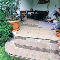 PAUL & ZBIG USŁUGI REMONTOWO - BUDOWLANE PAWEŁ FIJAŁKOWSKI & ZBIGNIEW ZALEWSKI S.C. - Mężczyzna w szarym ubraniu układa płytki na schodach tarasu, widoczne narzędzia, donice z kwiatami i drzewa w tle.