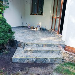 PAUL & ZBIG USŁUGI REMONTOWO - BUDOWLANE PAWEŁ FIJAŁKOWSKI & ZBIGNIEW ZALEWSKI S.C. - Wykończony taras z kamiennymi schodami, widoczne worki z zaprawą i narzędzia, oświetlenie girlandą żarówkową, mały krzew iglasty obok.