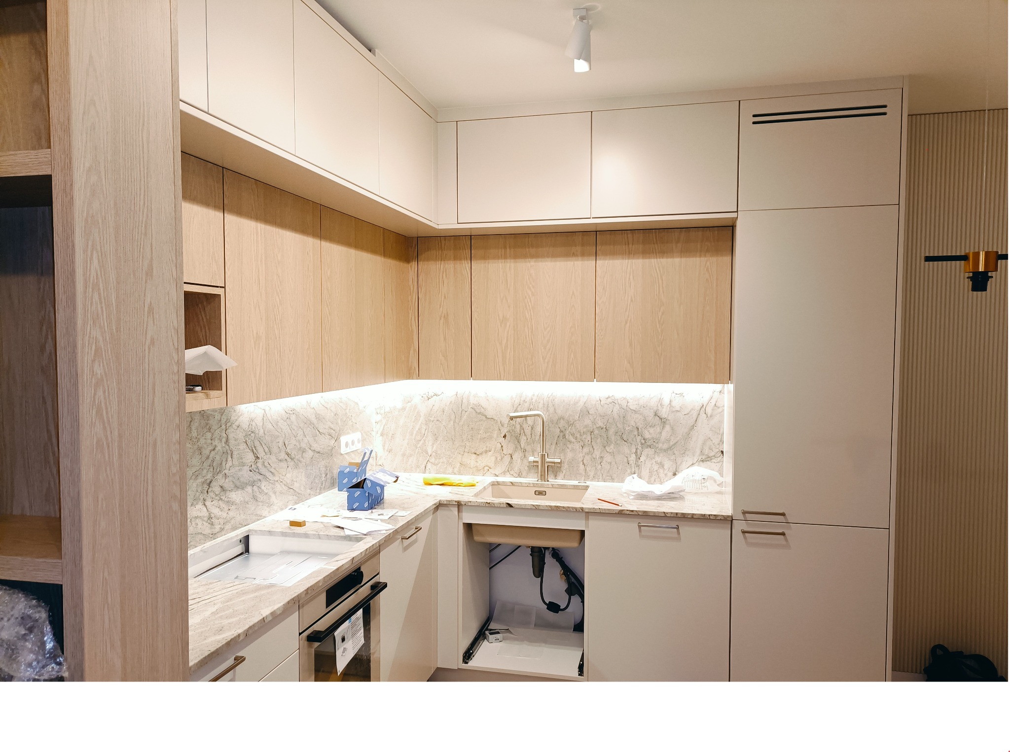 Kuchnia w trakcie montażu: jasne drewniane i białe szafki, marmurowy backsplash, widoczne elementy instalacji pod zlewem, kartonowe pudełka na blacie.