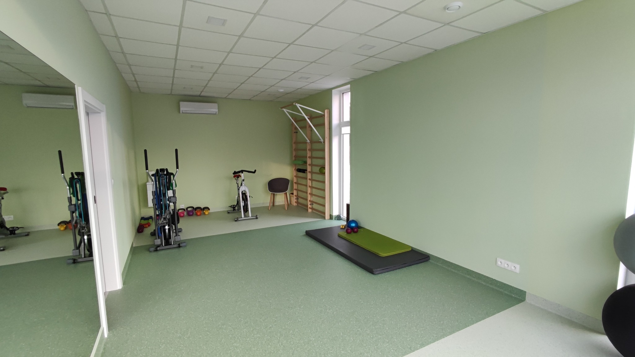 Jasnozielone ściany i podłoga w przestronnym pomieszczeniu ze sprzętem do ćwiczeń, w tym rower stacjonarny, drabinki gimnastyczne, mata do ćwiczeń i hantle. Widoczne gładkie ściany i sufit...