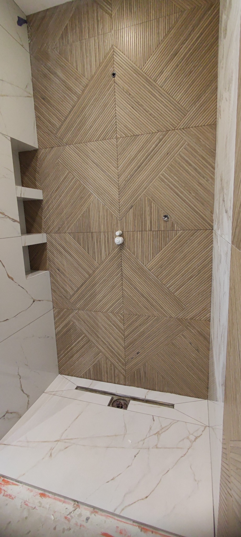 Wykończona łazienka z beżową ścianą z płytek ułożonych we wzór geometryczny i białą podłogą z odpływem liniowym.