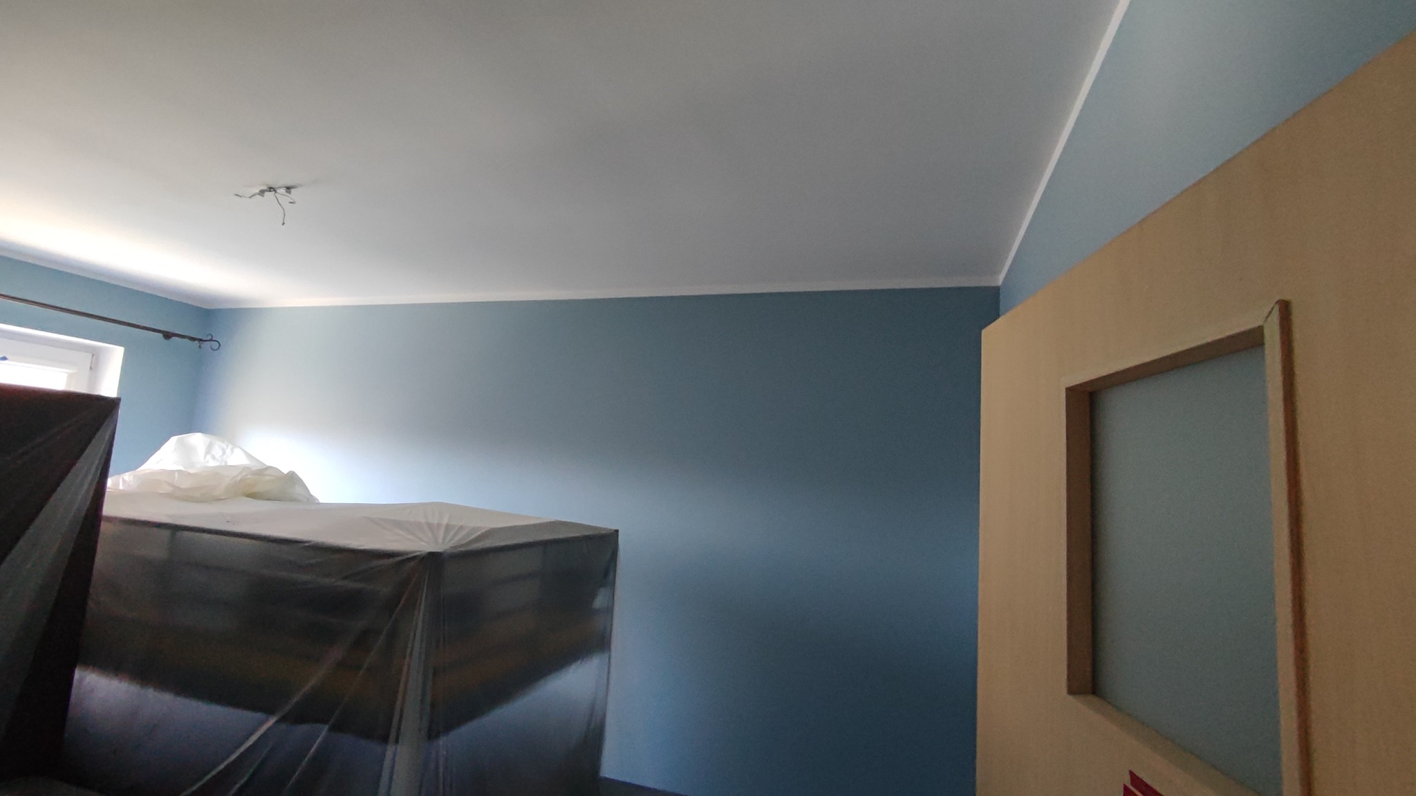 Pokój z pomalowanymi na niebiesko ścianami, biały sufit z wystającymi kablami, meble zabezpieczone folią ochronną, widoczny fragment futryny drzwi.