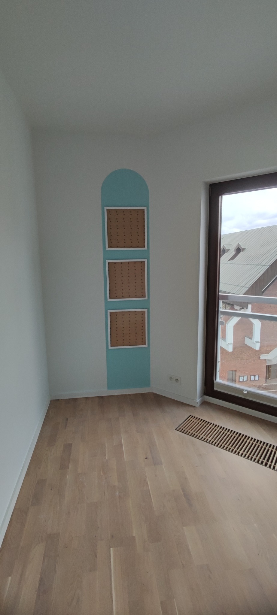Pusty pokój z drewnianą podłogą, białymi ścianami i pionowym, turkusowym panelem z trzema pustymi ramkami na ścianie, widok na budynki za oknem.