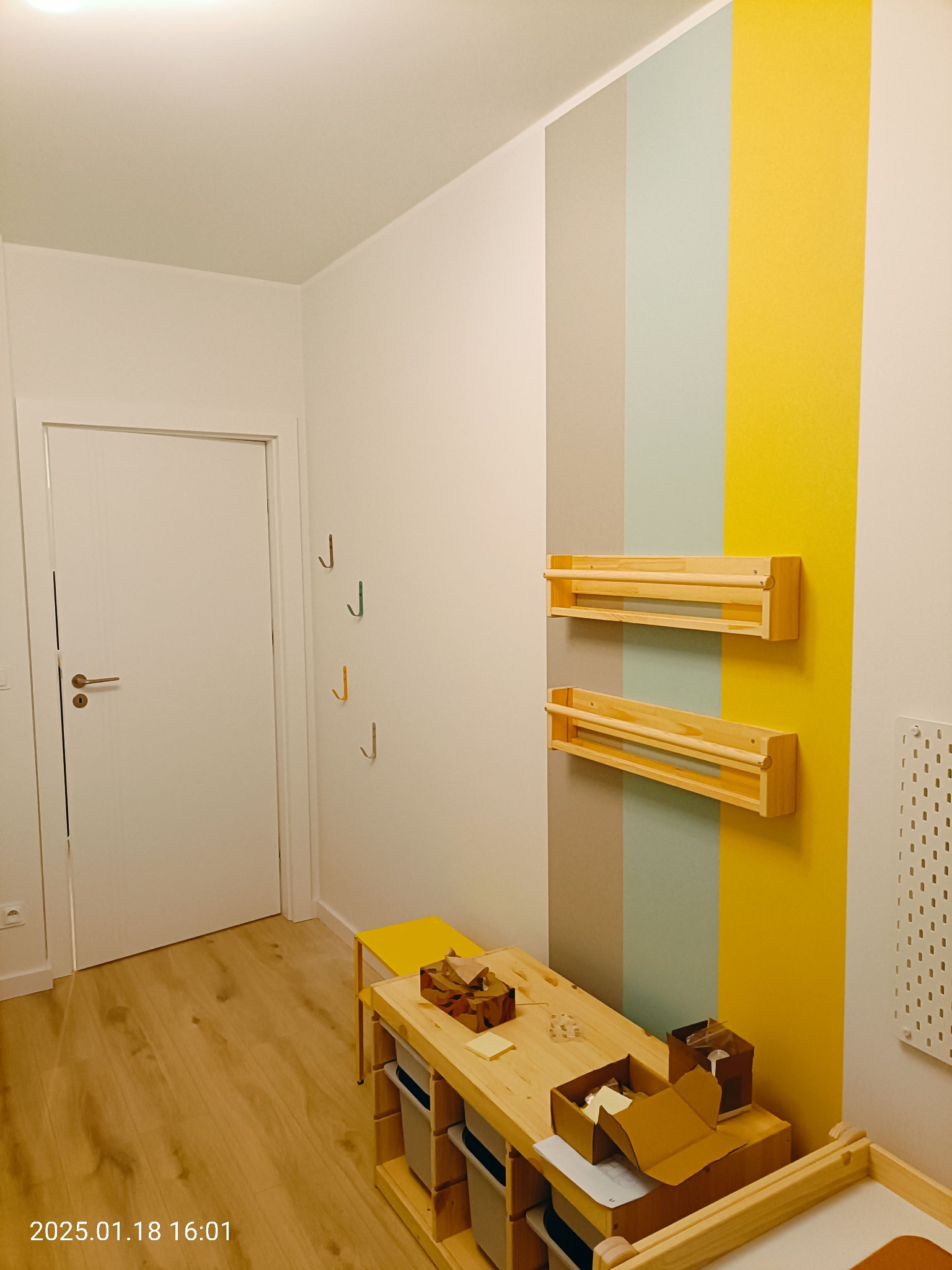 Pokój dziecięcy po odświeżeniu: ściana z pionowymi pasami w kolorach szarym, miętowym i żółtym, drewniane półki na ścianie, drewniany stół z pudełkami z zabawkami, podłoga laminowana imitująca...