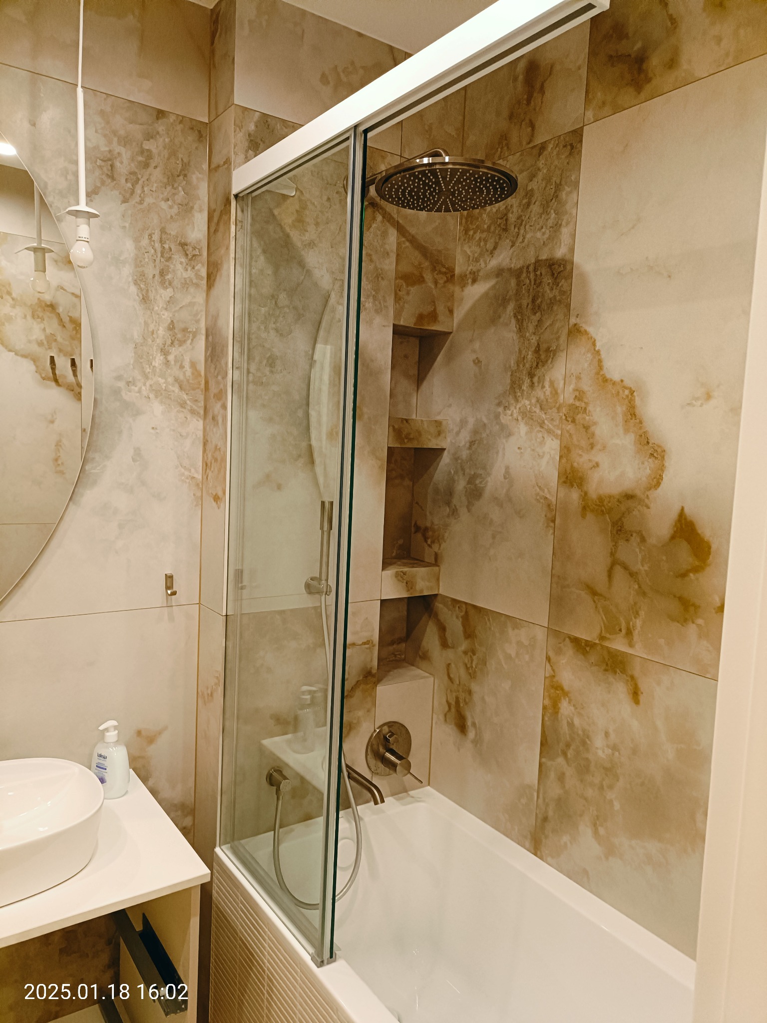 Wykończona łazienka z wanną z parawanem szklanym, deszczownicą i umywalką nablatową na szafce. Ściany wyłożone płytkami imitującymi marmur w odcieniach beżu i brązu.