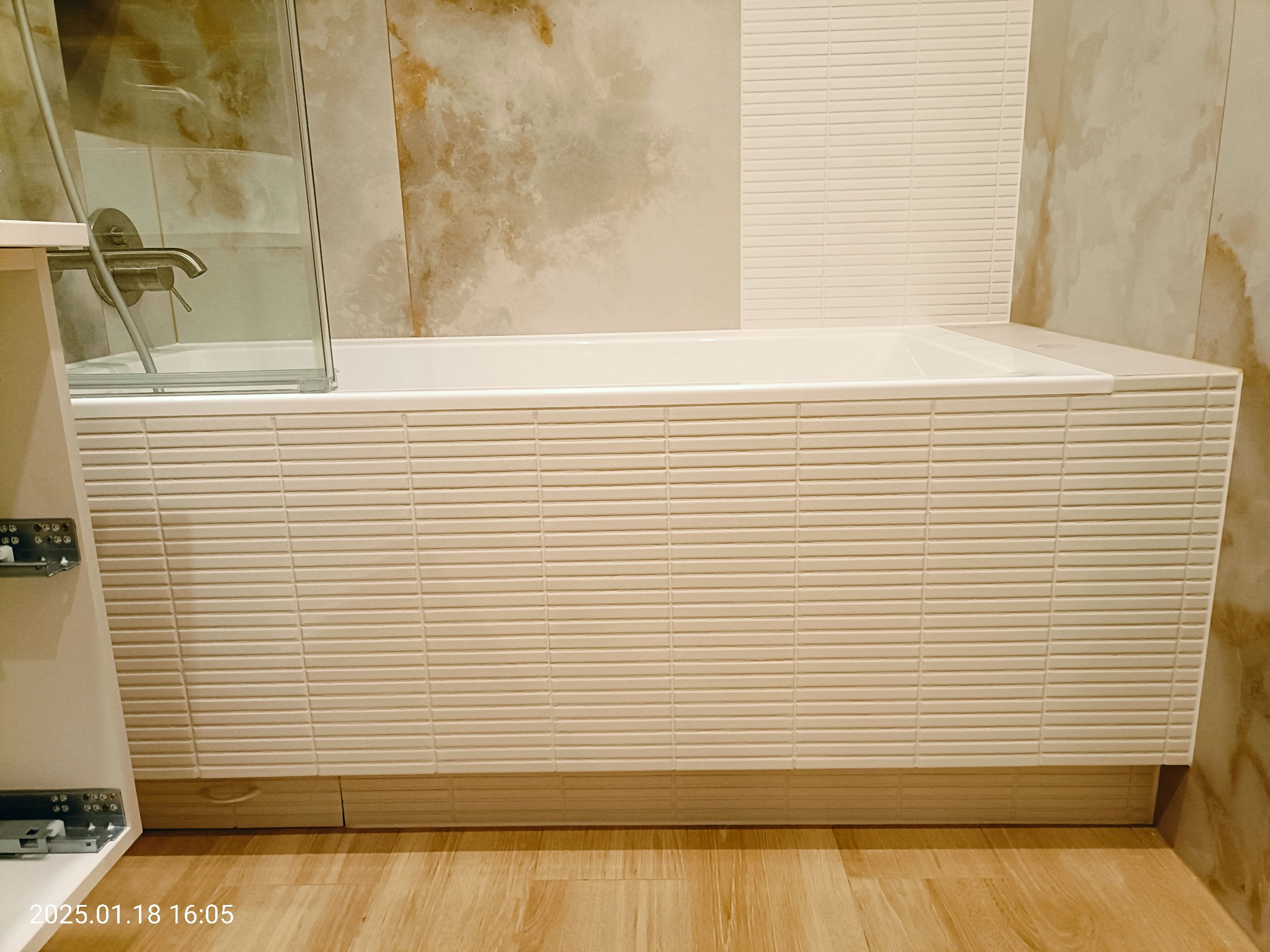 Wykończona wanna obłożona drobną, poziomą mozaiką w odcieniach beżu, z widoczną szklaną ścianką prysznicową i fragmentem szafki łazienkowej z systemem szuflad.