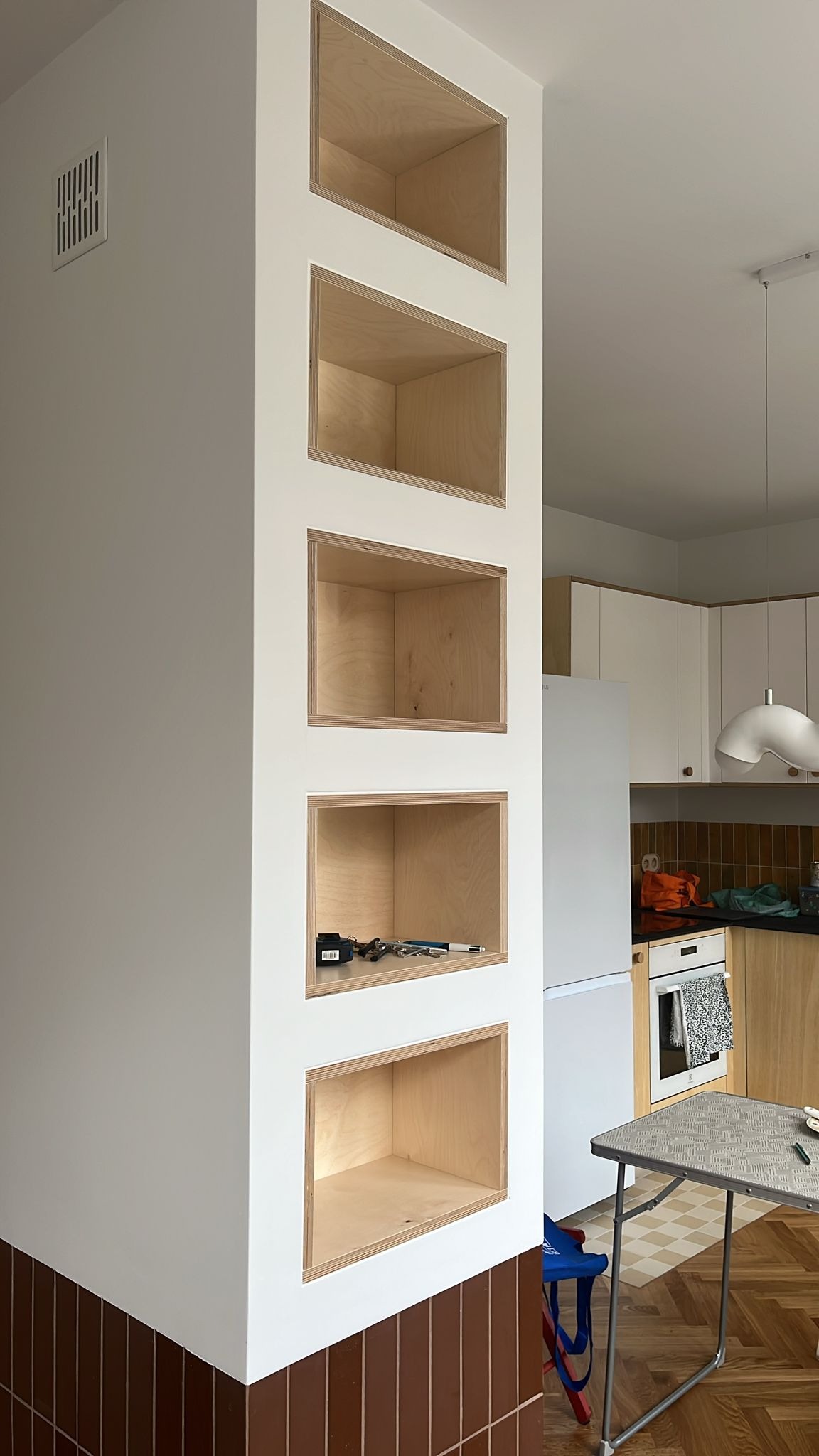 Wnęki z surowej sklejki w białej ścianie działowej, widoczne narzędzia w jednej z wnęk, brązowe płytki u dołu ściany, fragment kuchni w tle.