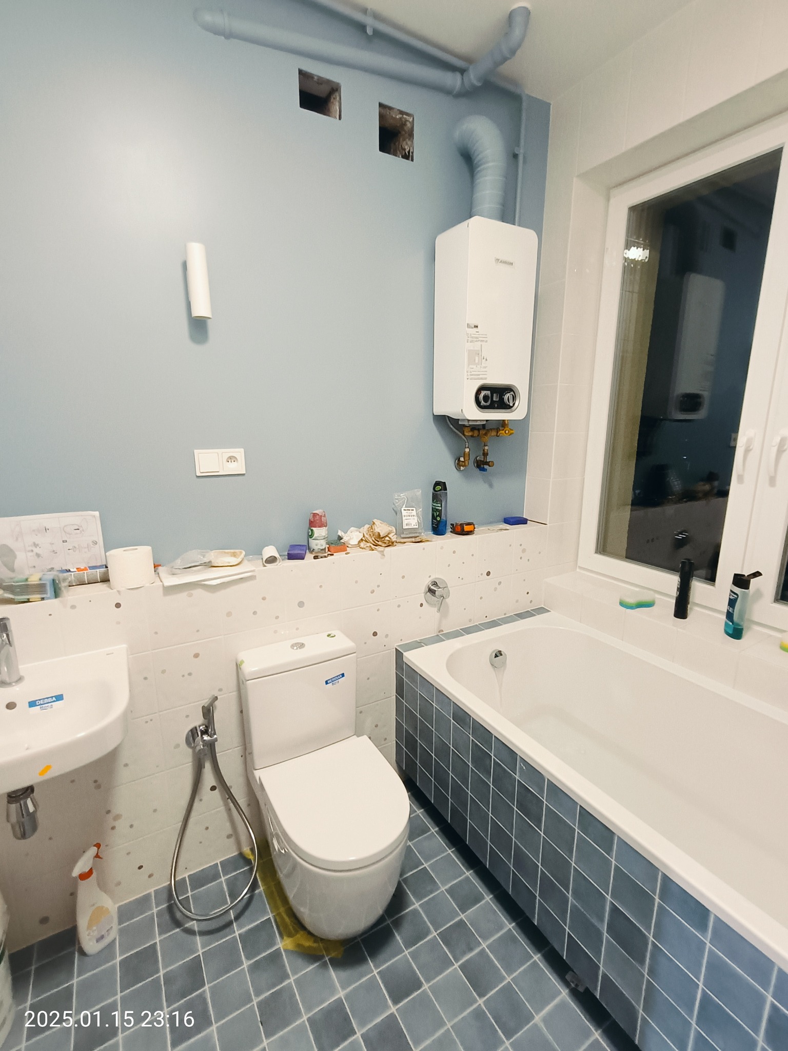 Łazienka w trakcie remontu: widoczny biały bojler nad wanną, umywalka, toaleta, niebieskie płytki na podłodze i częściowo na ścianach, ślady po pracach na ścianach, otwory w ścianie, rury, bałagan...