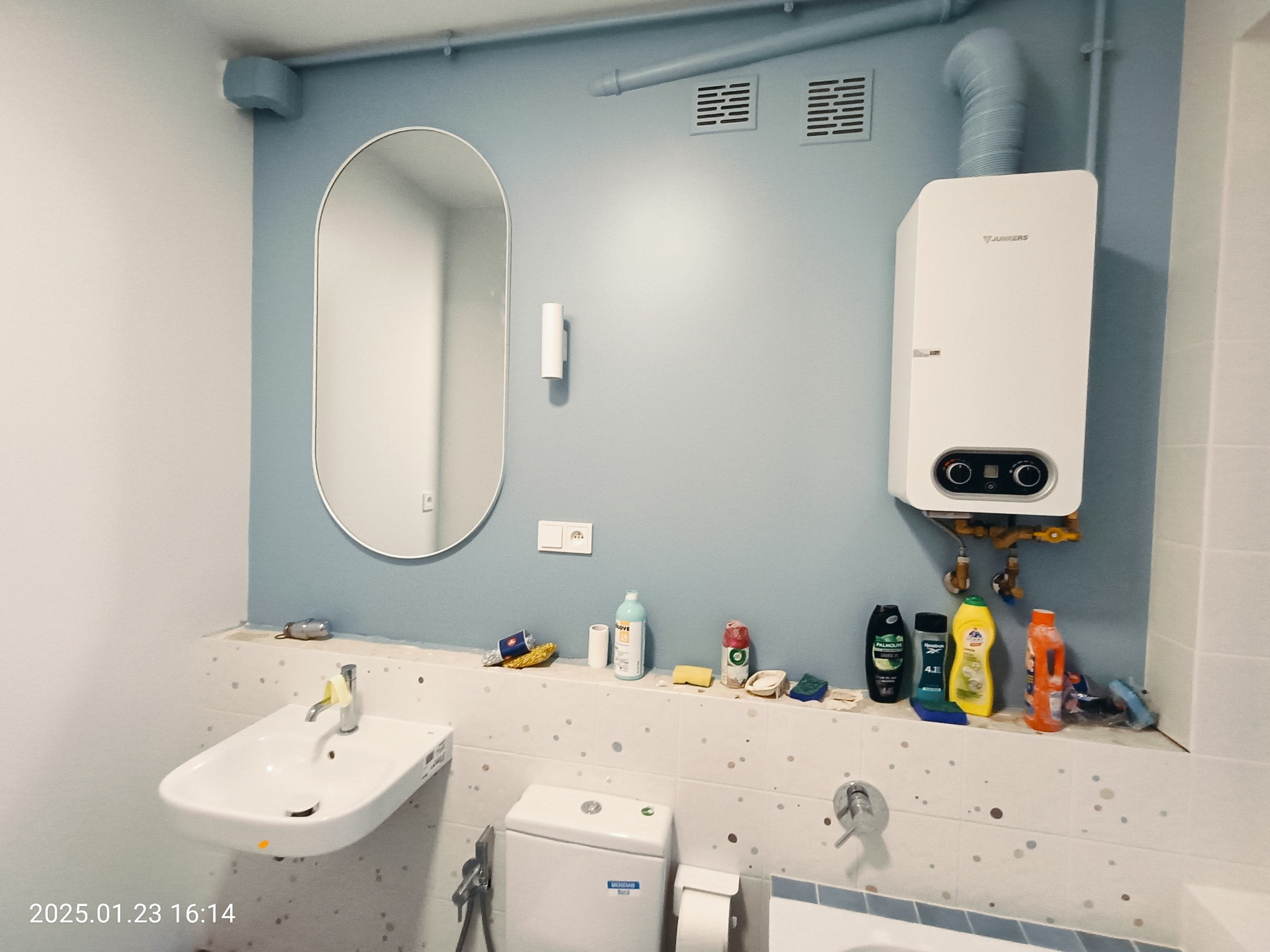 Niebieska łazienka z owalnym lustrem, białym zlewem, toaletą i bojlerem Junkers. Na półce nad toaletą stoją środki czystości i inne akcesoria łazienkowe. Widoczne rury i kratki wentylacyjne.