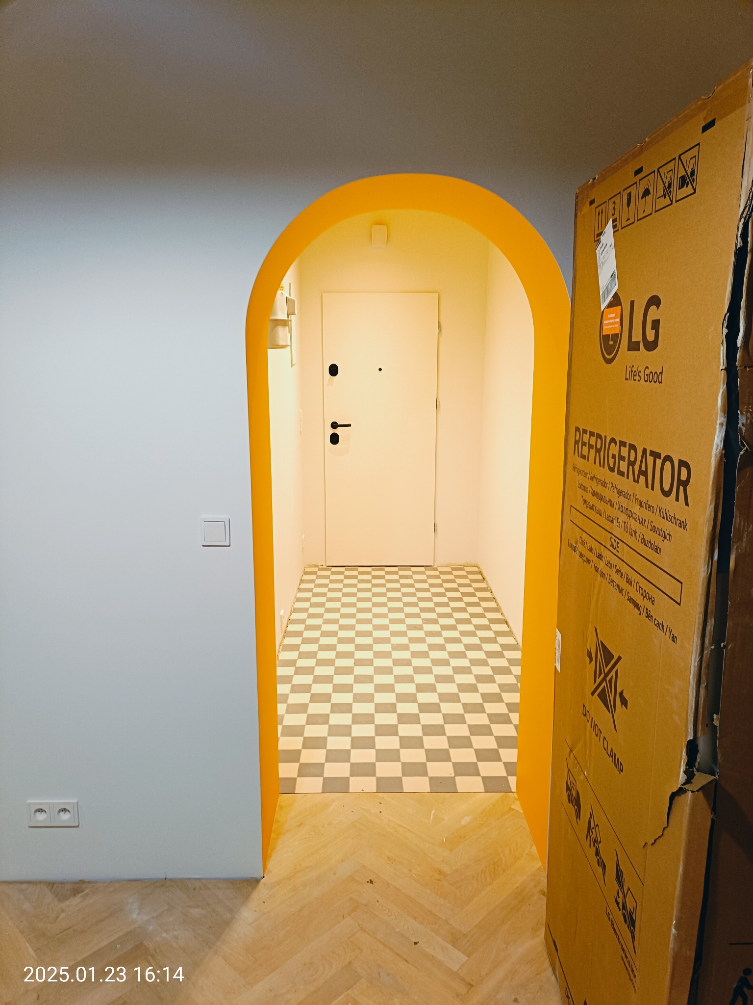 Korytarz z żółtym łukowym przejściem, podłoga w szachownicę, fragment podłogi wyłożony parkietem w jodełkę i karton po lodówce LG.