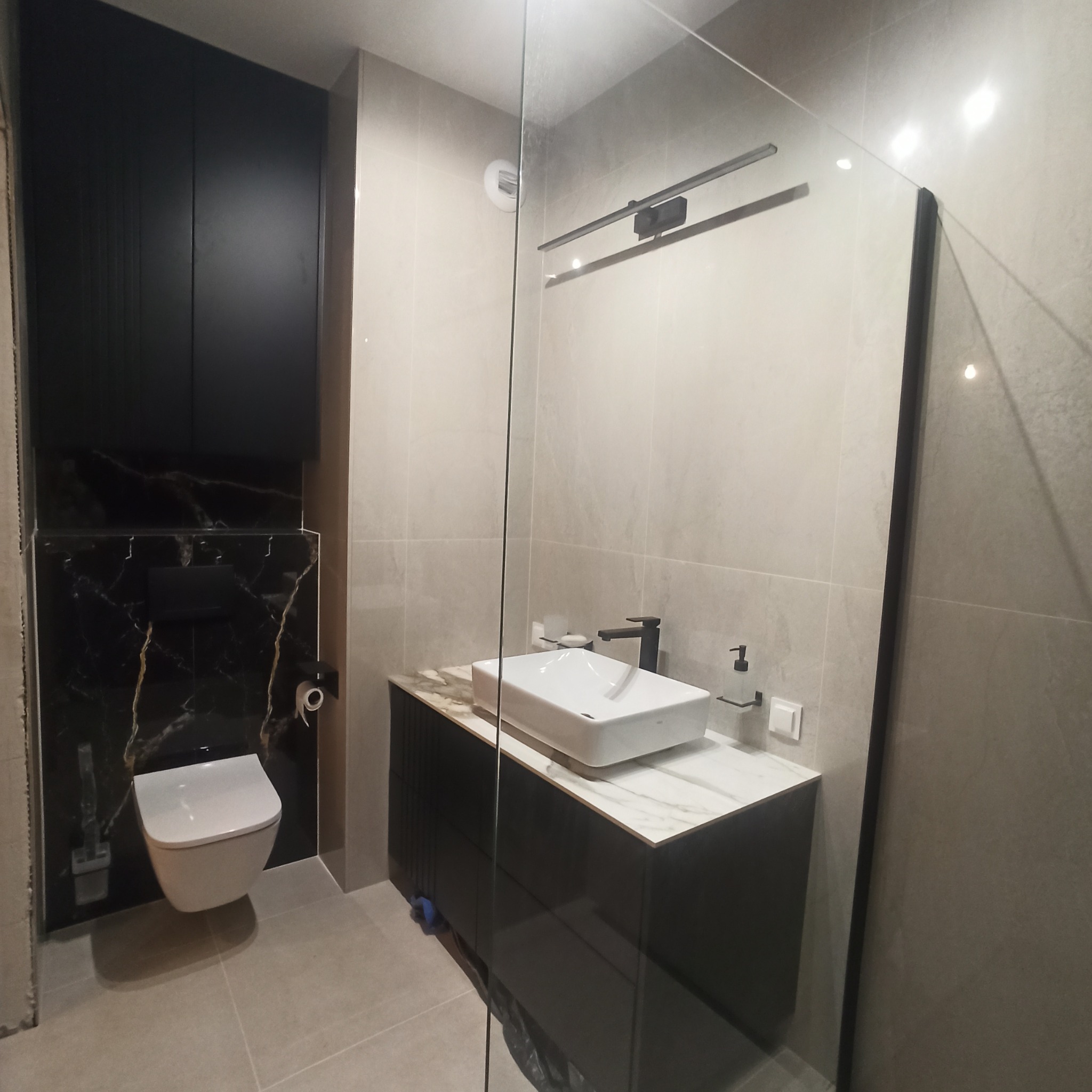 Nowoczesna łazienka z podwieszaną toaletą na czarnej marmurowej ścianie, prostokątną umywalką na szafce z blatem imitującym marmur i szklaną kabiną prysznicową.
