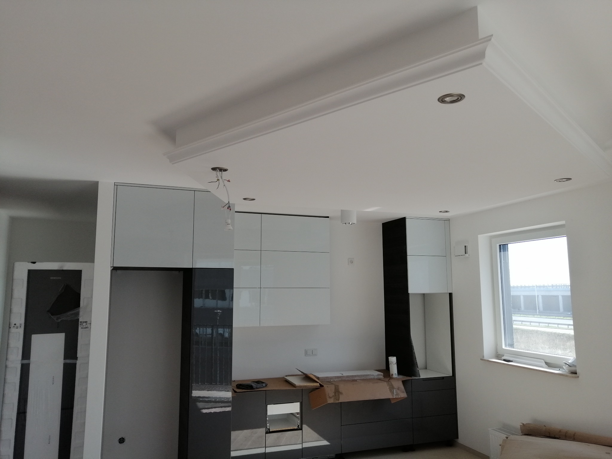 Montaż nowoczesnej kuchni z białymi i grafitowymi frontami szafek, widoczne elementy wykończenia sufitu z dekoracyjną listwą i oświetleniem punktowym, okno z widokiem na wiadukt.