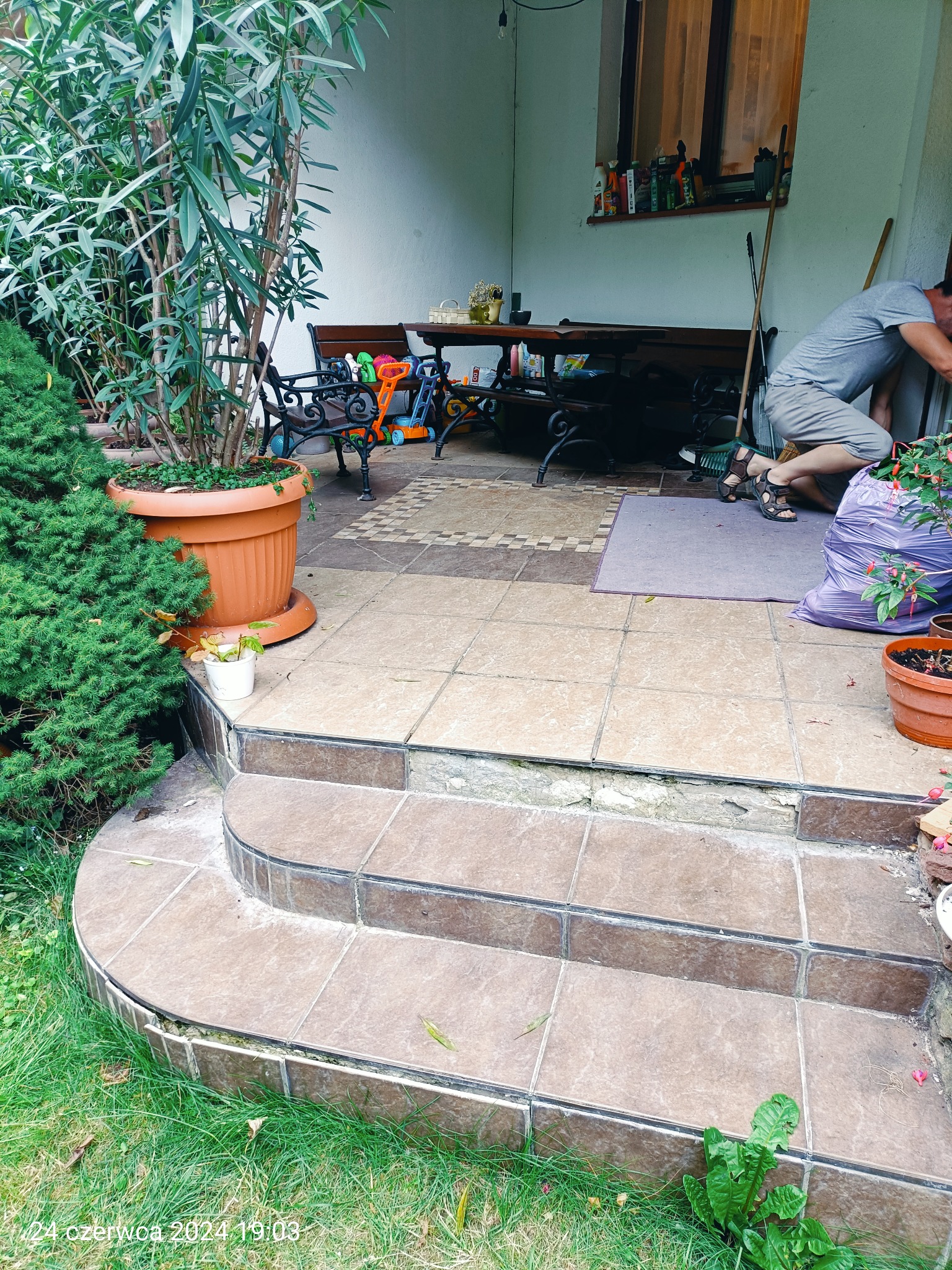 Mężczyzna w szarym ubraniu układa płytki na schodach tarasu, widoczne narzędzia, donice z kwiatami i drzewa w tle.