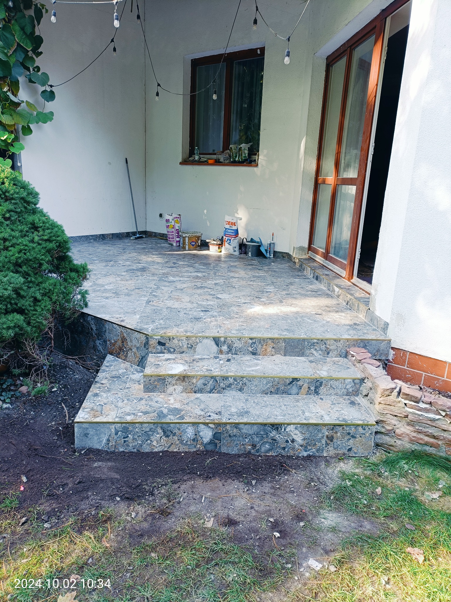 Wykończony taras z kamiennymi schodami, widoczne worki z zaprawą i narzędzia, oświetlenie girlandą żarówkową, mały krzew iglasty obok.