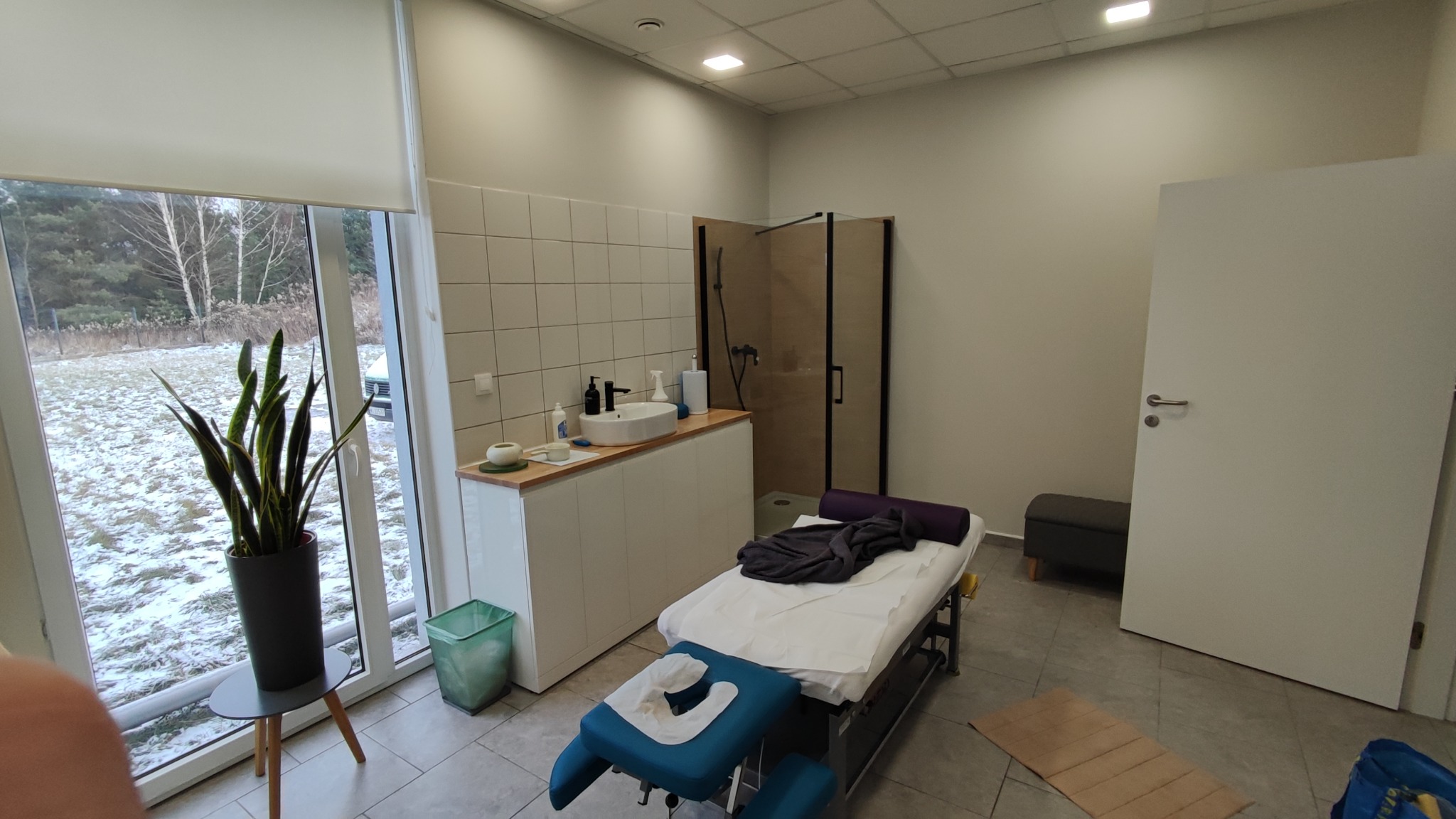 Jasne wnętrze gabinetu z łazienką, wyposażone w kozetkę z niebieskim zagłówkiem, umywalkę z białą szafką, przeszkloną kabinę prysznicową oraz dużą roślinę doniczkową przy oknie z widokiem...