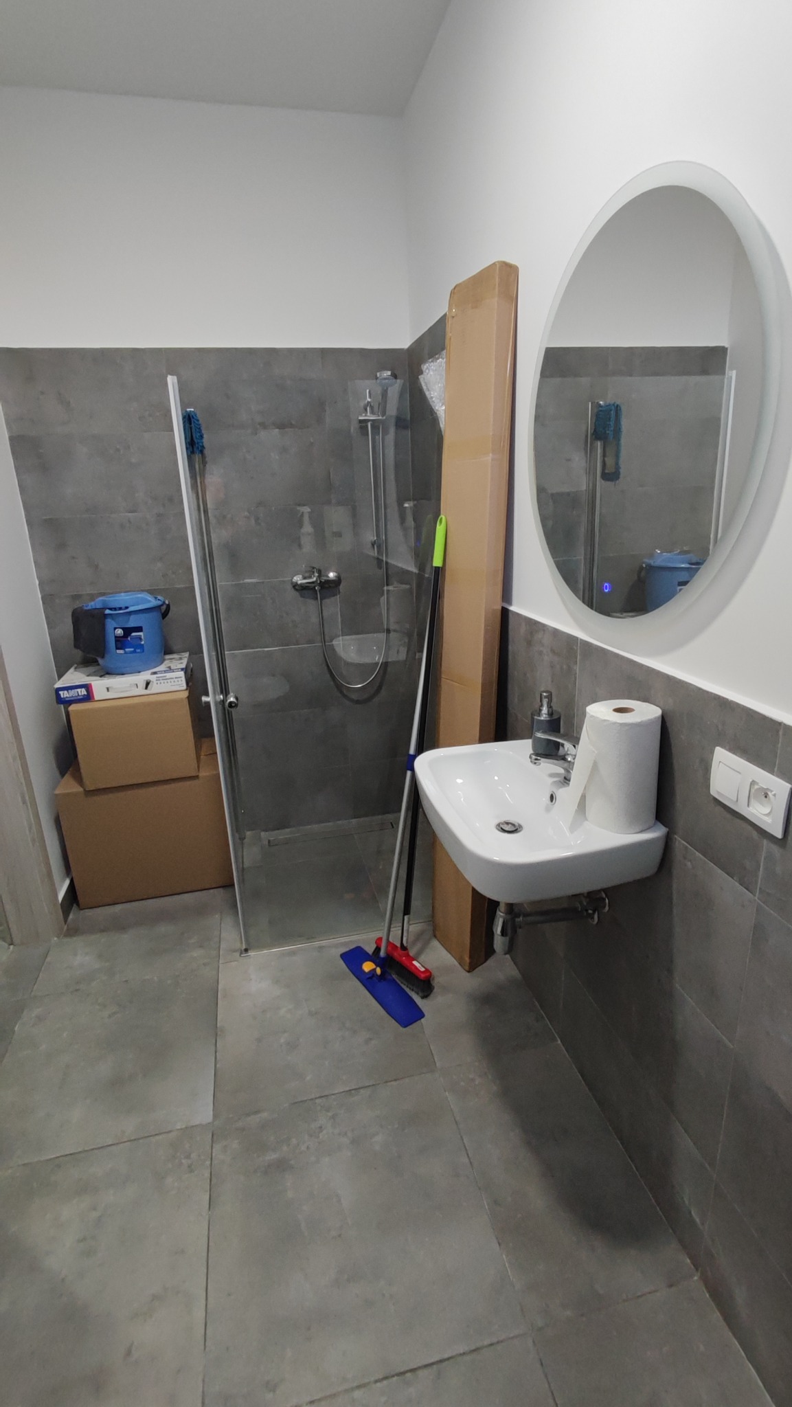 Łazienka w trakcie prac wykończeniowych: kabina prysznicowa ze szklanymi drzwiami, umywalka z armaturą, owalne lustro, kartony i akcesoria malarskie, szara płytka na ścianach i podłodze.