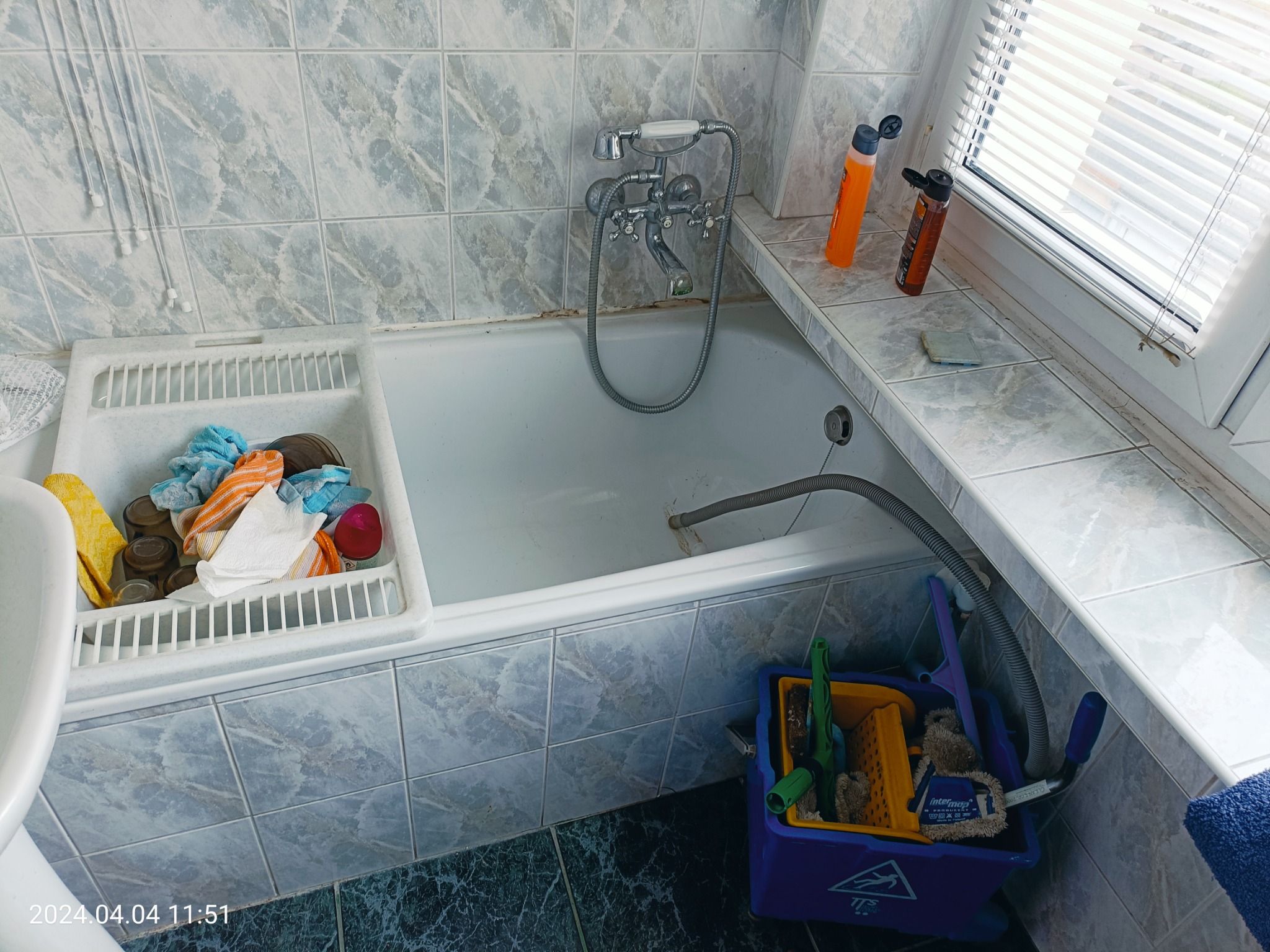 Niezbyt czysta wanna z baterią, przy oknie butelki z płynami, obok wanny wiadro z narzędziami i akcesoriami do sprzątania, widoczne ślady użytkowania.