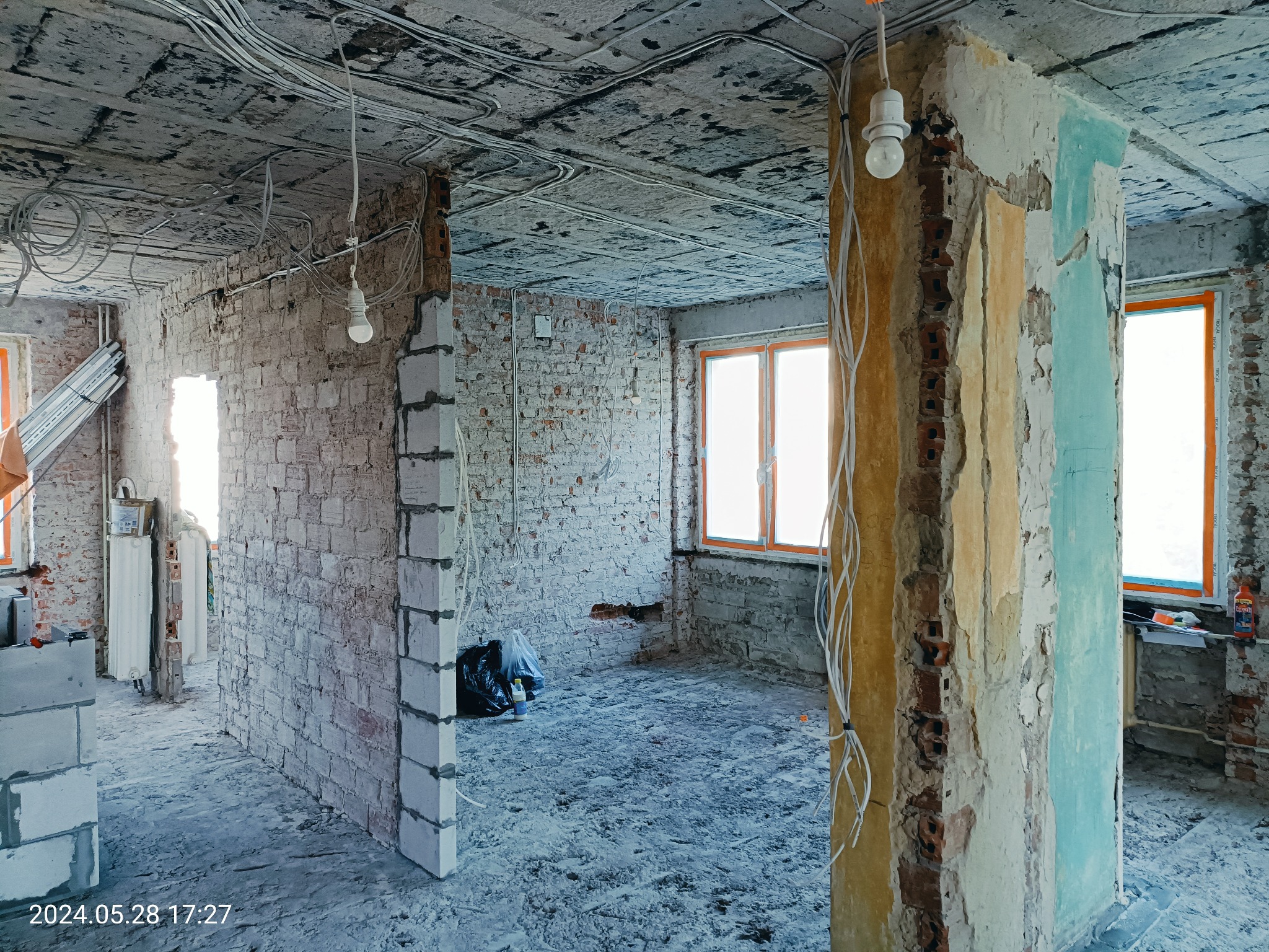 Wnętrze w trakcie remontu: odsłonięte cegły, surowy betonowy sufit z widoczną instalacją elektryczną, kolumna z resztkami farby i tynku w odcieniach beżu i turkusu, tymczasowe oświetlenie żarówką.