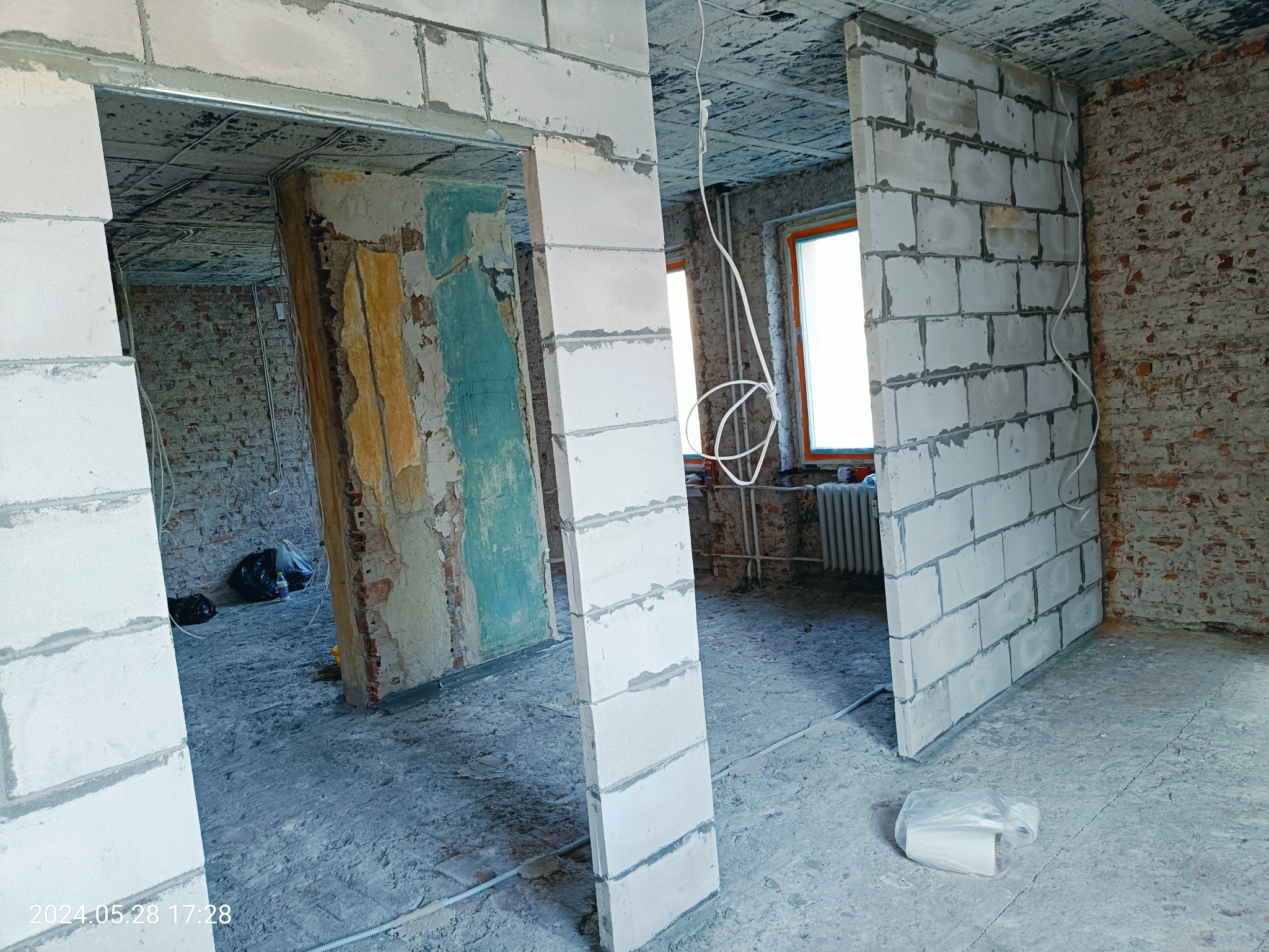 Wnętrze w trakcie budowy ścian działowych z bloczków, widoczne surowe ściany z cegły i fragmenty starej farby, przewody elektryczne zwisające z sufitu, podłoga pokryta pyłem budowlanym, foliowy...