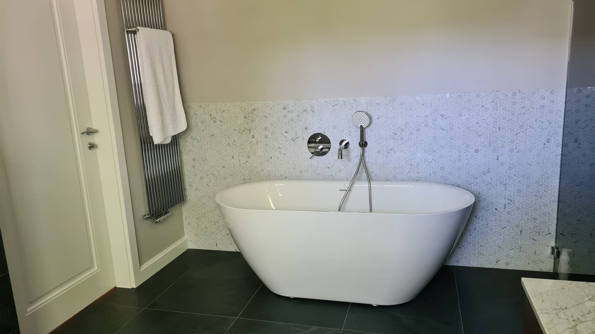 Nowoczesna łazienka z wolnostojącą białą wanną, chromowaną armaturą i grzejnikiem drabinkowym z powieszonym białym ręcznikiem, wykończona mozaiką na ścianie.