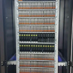 Pełna szafa serwerowa z dużą ilością dysków twardych w obudowach z pomarańczowymi elementami, widoczna z bliska w ciemnym pomieszczeniu.