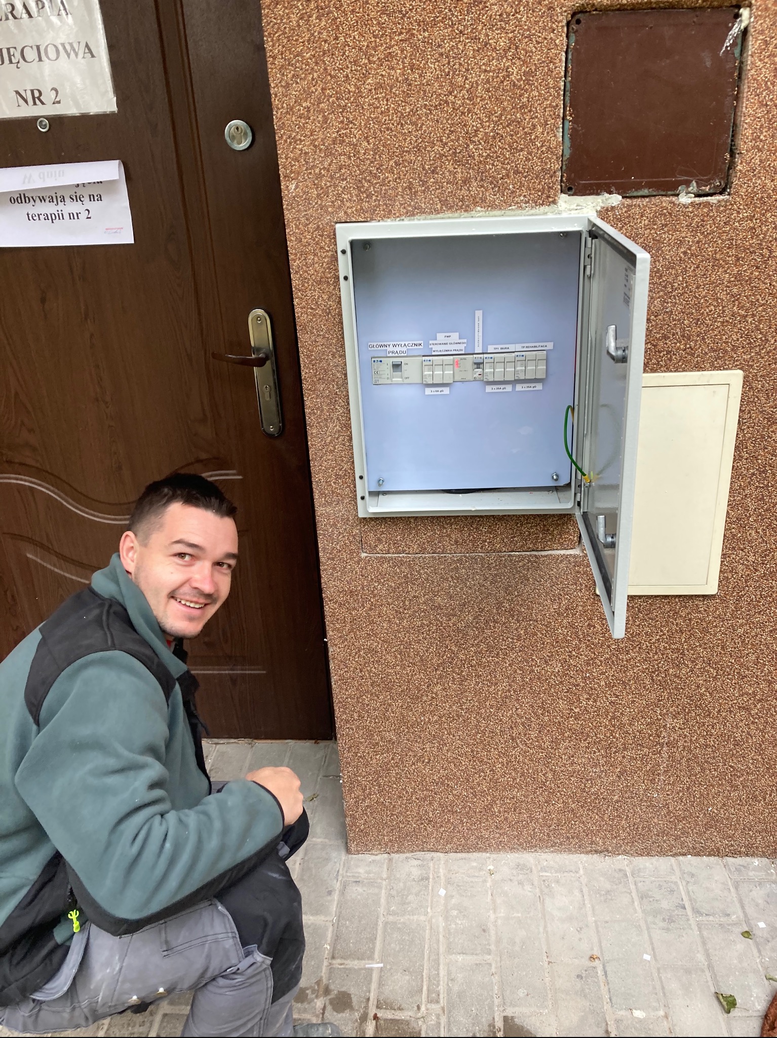 Uśmiechnięty mężczyzna w roboczym ubraniu kuca obok otwartej skrzynki z bezpiecznikami przy ścianie budynku z brązową elewacją, obok brązowych drzwi z kartką informacyjną.