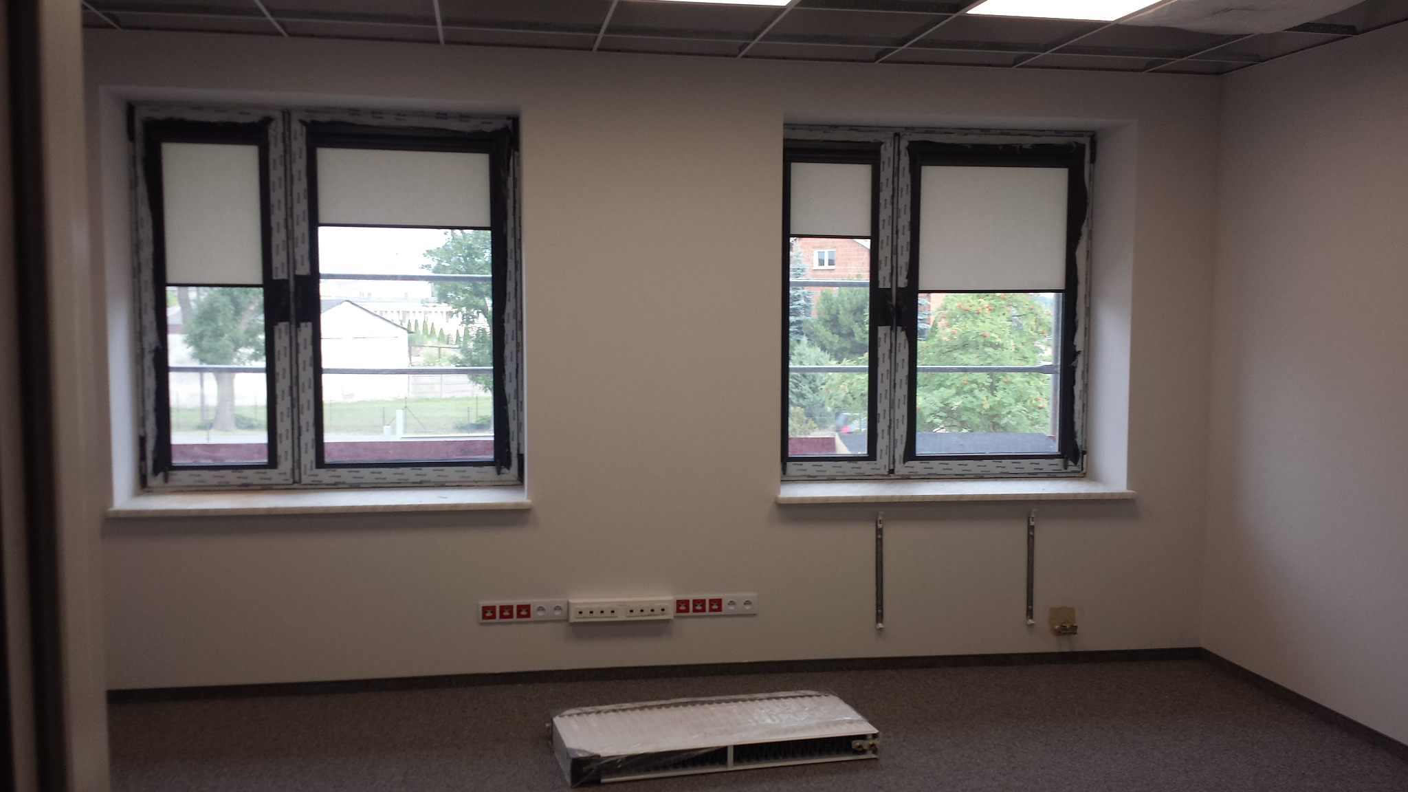 Puste pomieszczenie biurowe z dwoma oknami z roletami, grzejnikiem pod oknem i puszką instalacyjną na ścianie, przygotowane do instalacji elektrycznej.