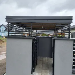 Nowoczesna wiata garażowa z metalową konstrukcją i betonowymi słupkami w szarej kolorystyce, widok z przodu, na tle domów jednorodzinnych i pochmurnego nieba.