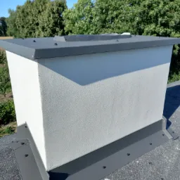 Otynkowana podstawa komina na dachu krytym papą, z szarymi obróbkami blacharskimi mocowanymi na śruby.