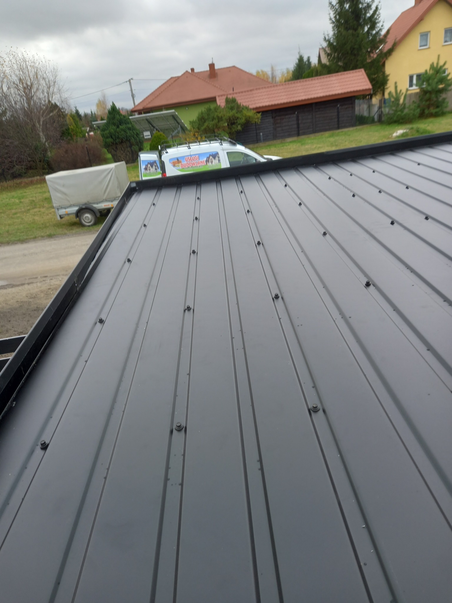 Szary, blaszany dach z widocznymi śrubami mocującymi, w tle zaparkowany biały bus z reklamą firmy budowlanej i przyczepa, pochmurne niebo.
