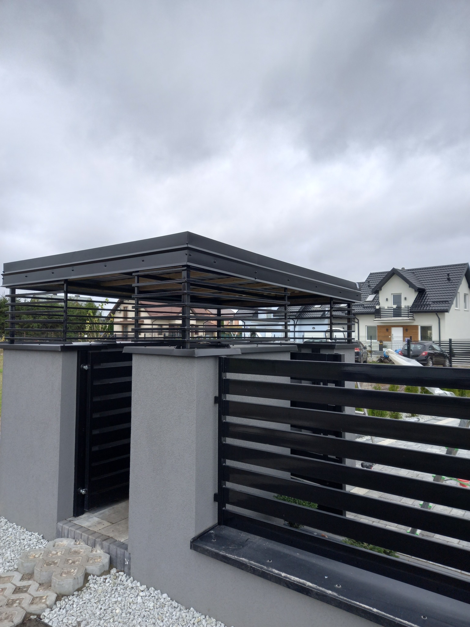 Nowoczesna, czarna konstrukcja zadaszenia tarasu lub wiaty, połączona z ogrodzeniem posesji, na tle szarego nieba i zabudowy jednorodzinnej.
