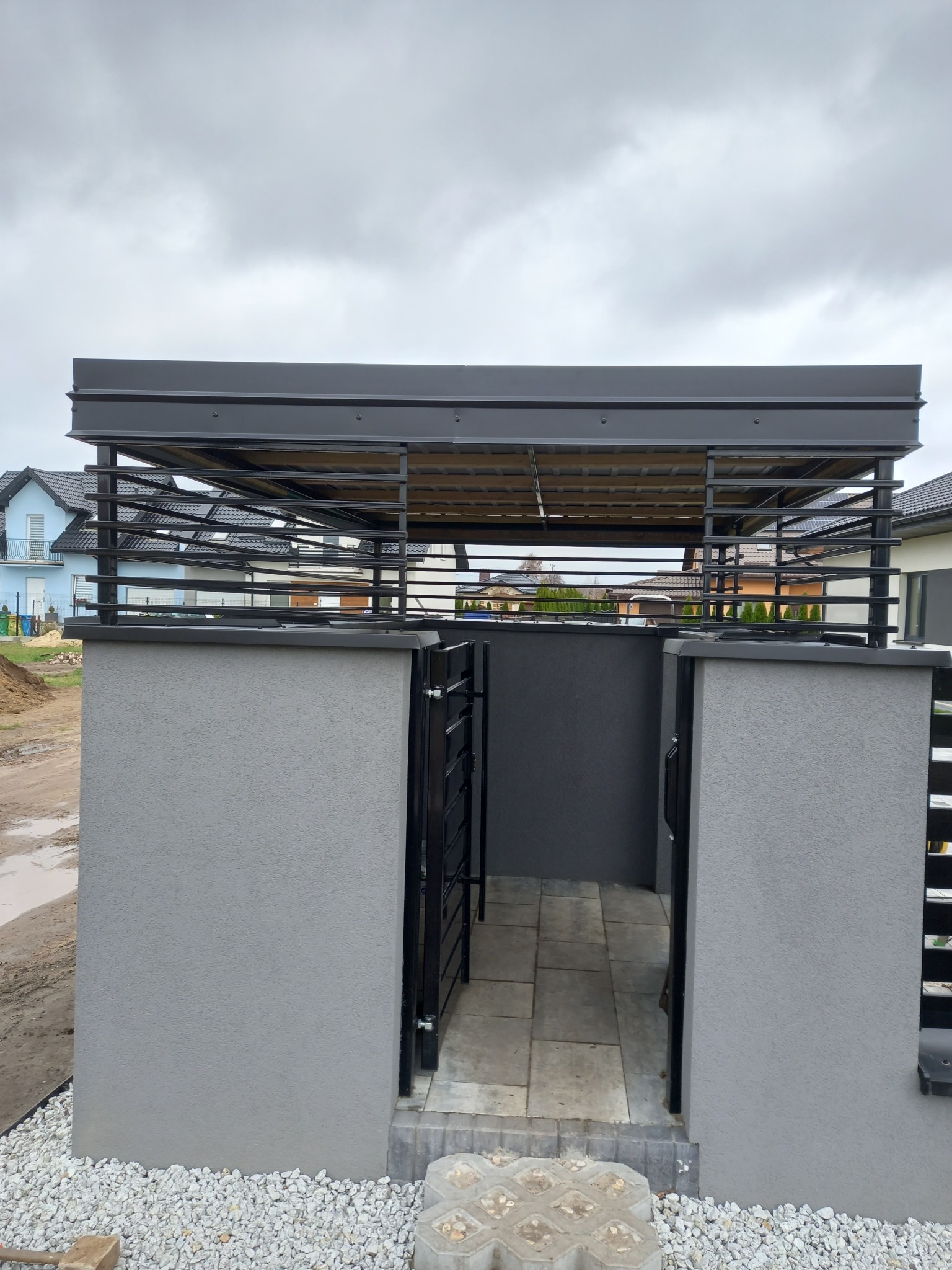 Nowoczesna wiata garażowa z metalową konstrukcją i betonowymi słupkami w szarej kolorystyce, widok z przodu, na tle domów jednorodzinnych i pochmurnego nieba.