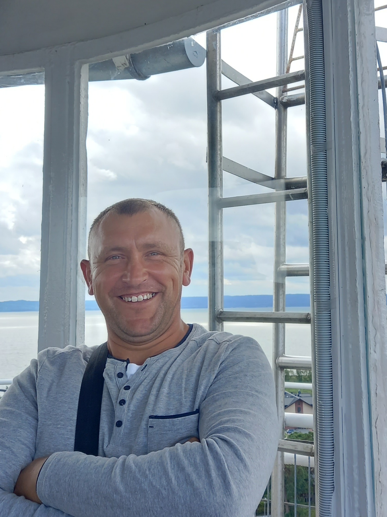 Uśmiechnięty mężczyzna w szarej koszulce na tle metalowej konstrukcji i widoku na jezioro zza okna.