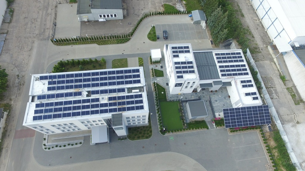 Widok z lotu ptaka na dwa budynki z panelami słonecznymi na dachach, otoczone zagospodarowanym terenem z trawnikami i drzewami, przylegający parking z zaparkowanym samochodem.