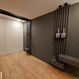 Nowoczesna szafa wnękowa z białymi frontami, drewniana podłoga, szara ściana z dekoracyjnymi panelami i oświetleniem.