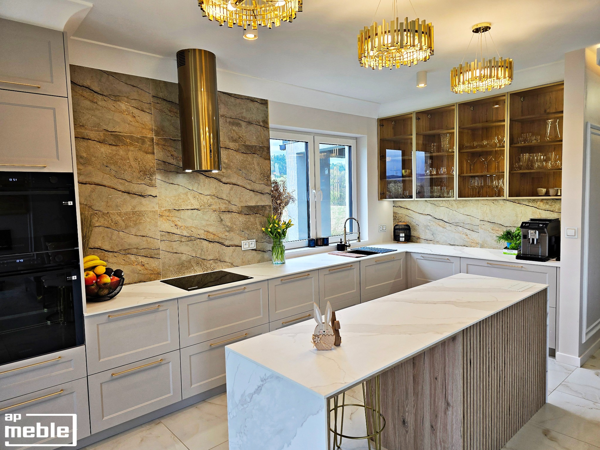 Elegancka kuchnia w odcieniach szarości z wyspą i złotymi akcentami. Marmurowe płytki na ścianie, złoty okap i lampy nadają luksusowy charakter wnętrzu.