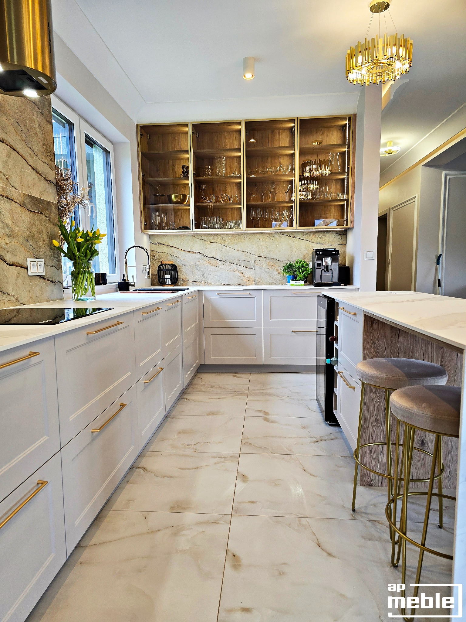 Elegancka kuchnia z białymi frontami, złotymi uchwytami i marmurowym blatem. Na wyspie dwa hokery ze złotymi nogami. Nad blatem szafki z przeszkleniami.