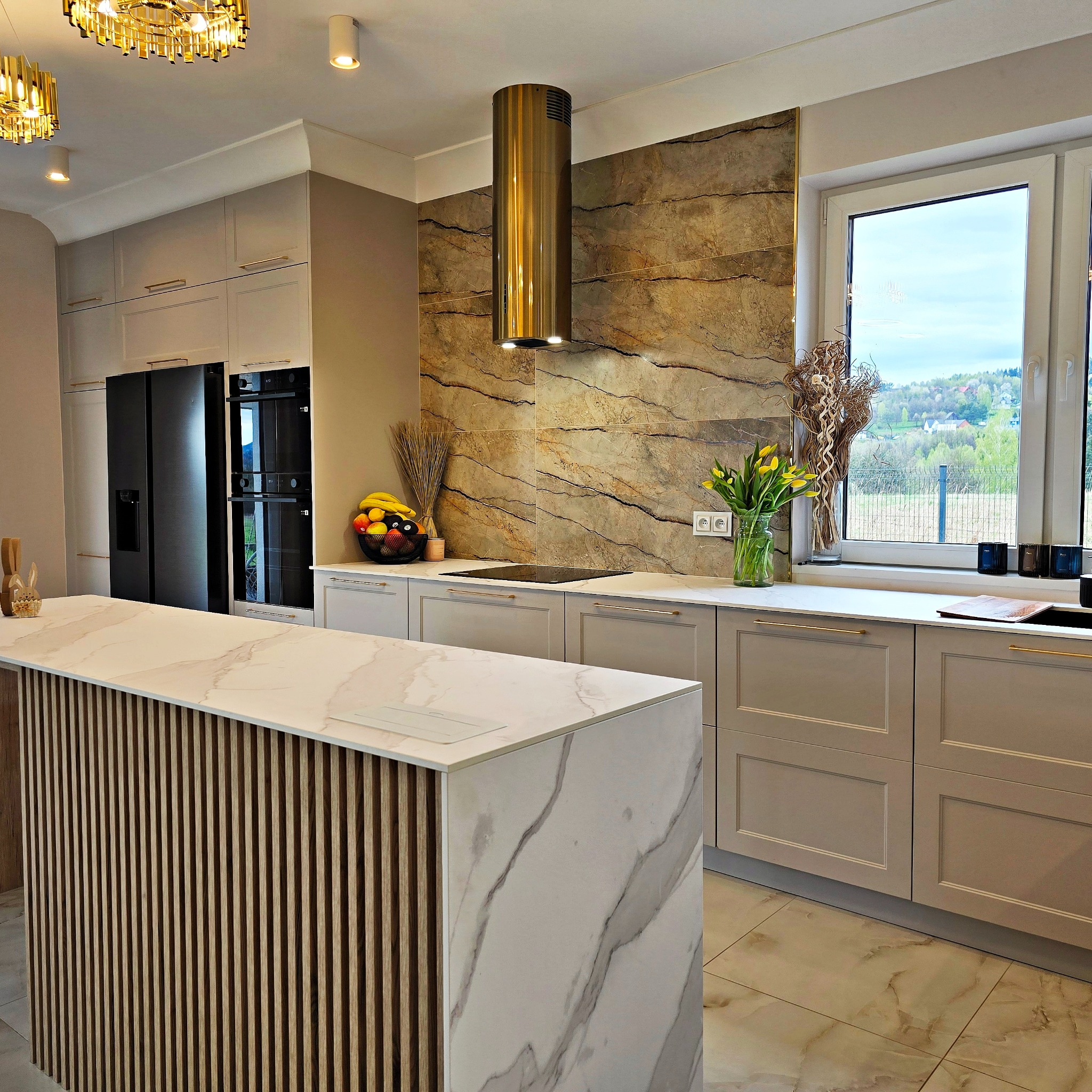 Nowoczesna kuchnia z wyspą: beżowe szafki z złotymi uchwytami, marmurowy blat, złoty okap cylindryczny i widok na zieleń za oknem.