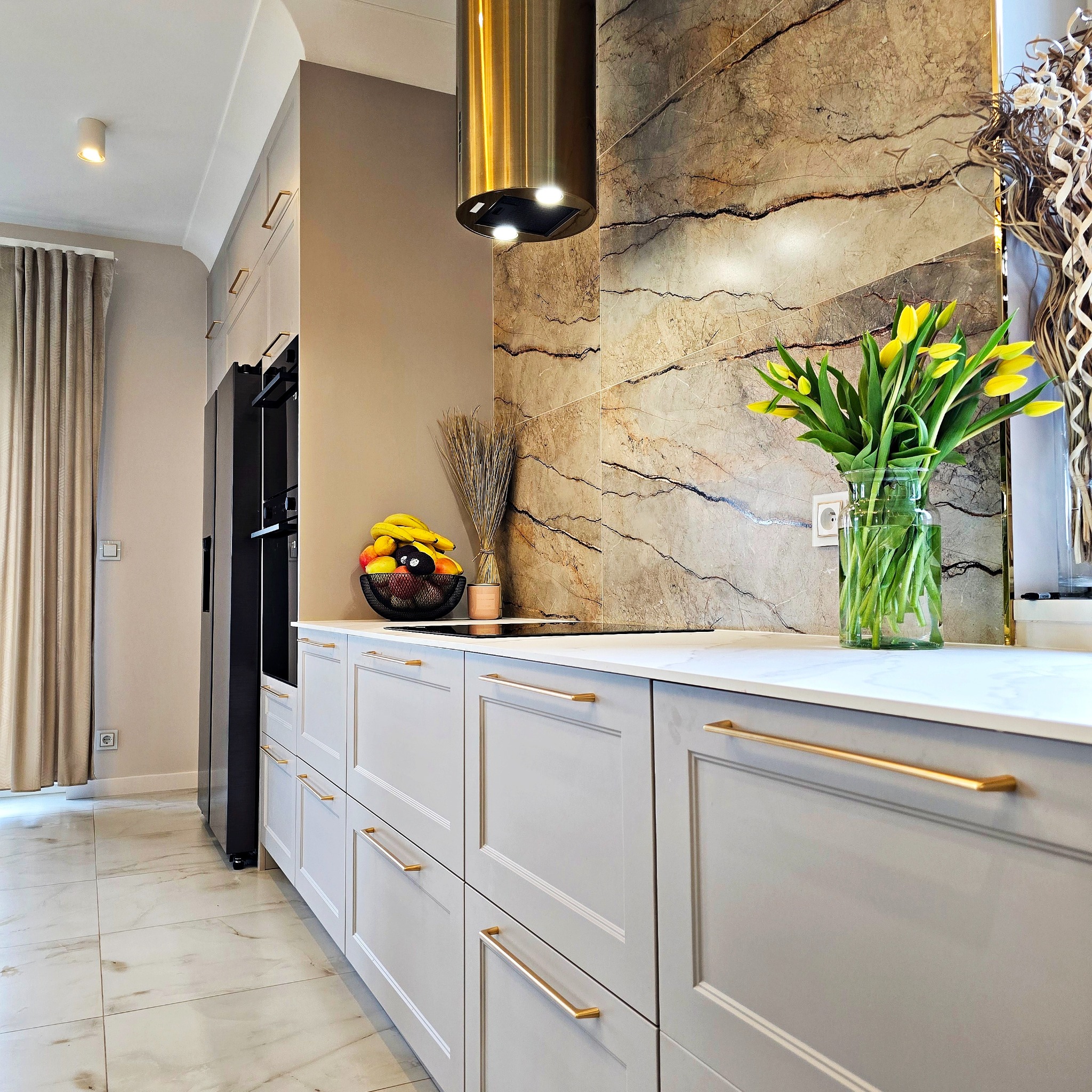 Elegancka kuchnia z szarymi szafkami, złotymi uchwytami i marmurowym blatem. Na blacie owoce w koszyku i żółte tulipany w szklanym wazonie. Nowoczesny okap.