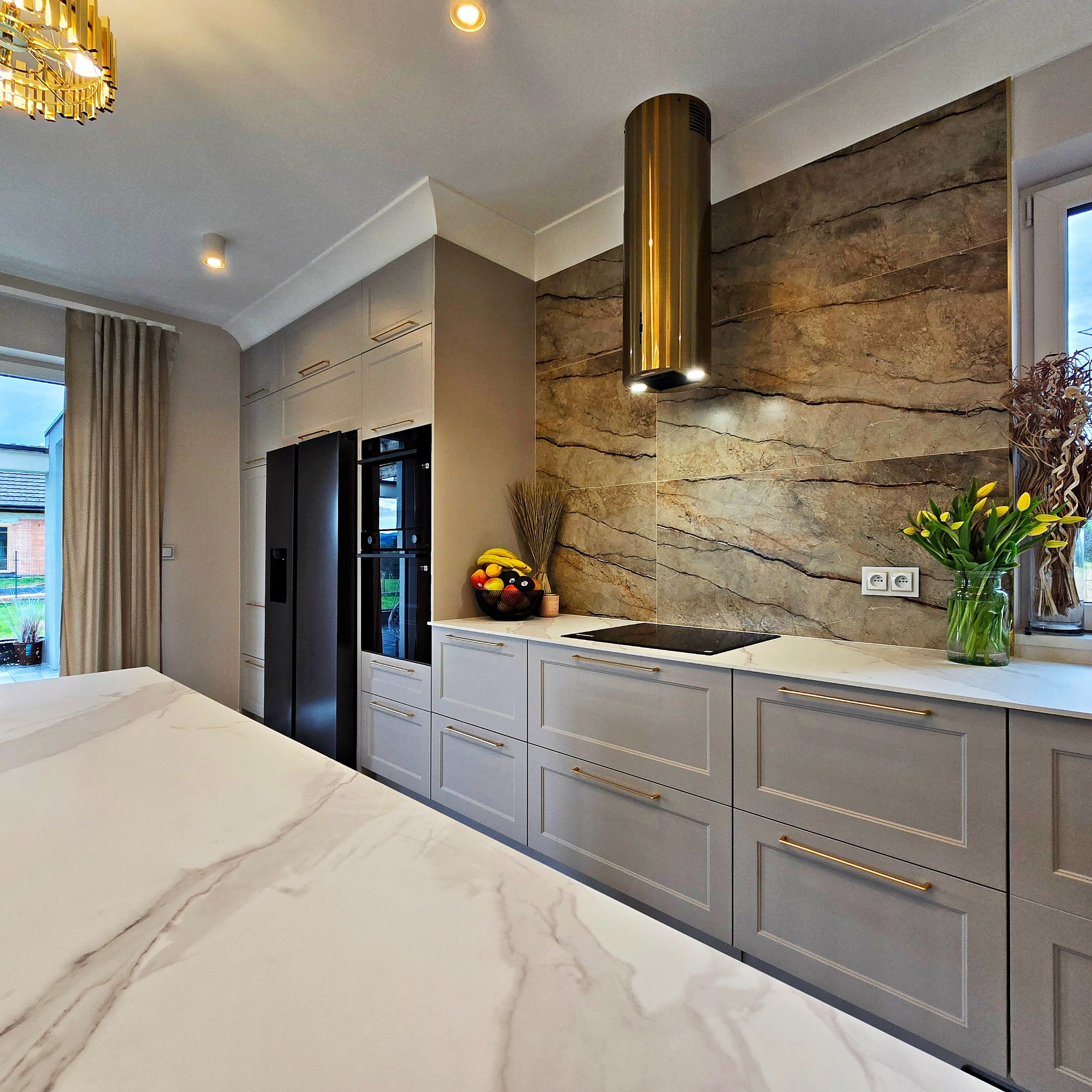 Nowoczesna kuchnia z szarymi szafkami, złotymi uchwytami i marmurowym blatem. Na ścianie dekoracyjny panel imitujący marmur oraz złoty okap.