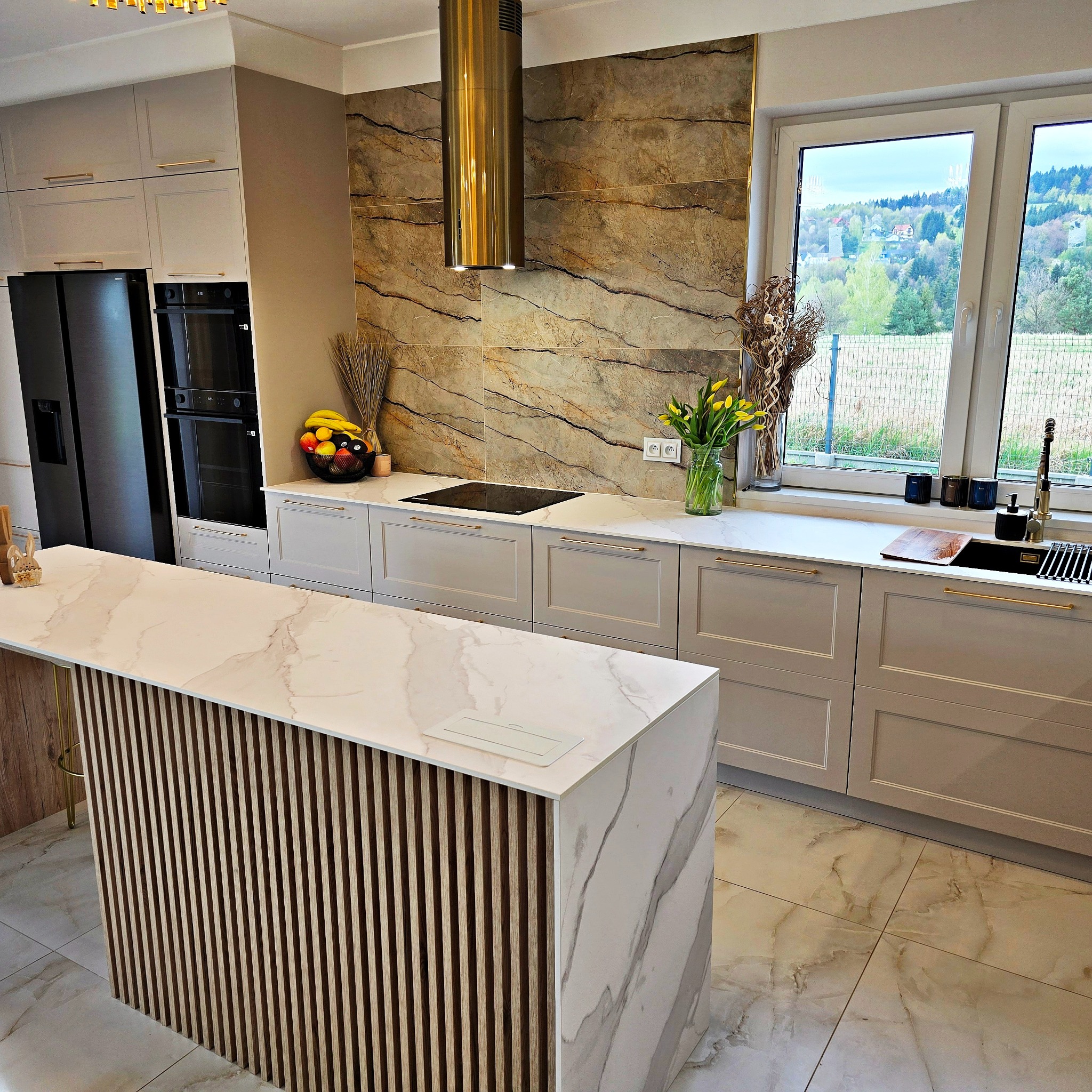 Elegancka kuchnia z wyspą o marmurowym blacie i drewnianymi lamelami, złoty okap cylindryczny, beżowe szafki z długimi uchwytami i widok na zielone wzgórza za oknem.