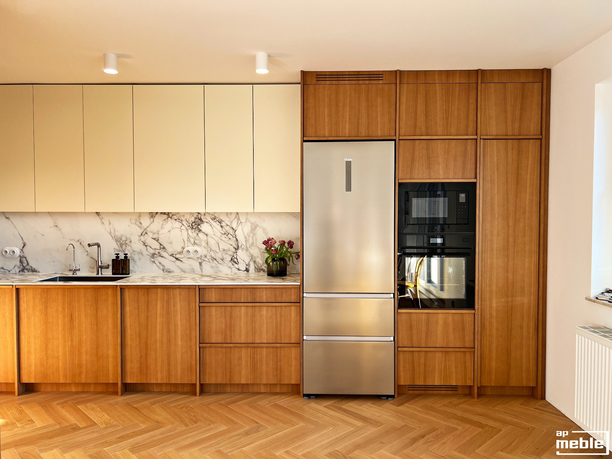Nowoczesna kuchnia z drewnianymi frontami, marmurowym blatem i srebrną lodówką. Parkiet w jodełkę. Ujęcie z niskiego kąta podkreśla przestronność wnętrza.