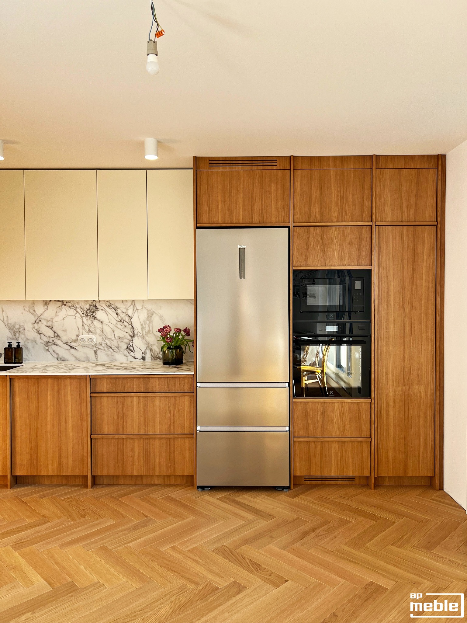 Nowoczesna kuchnia z drewnianymi frontami, marmurowym blatem i srebrną lodówką. Parkiet w jodełkę. Widoczne oświetlenie sufitowe i kwiaty w wazonie.