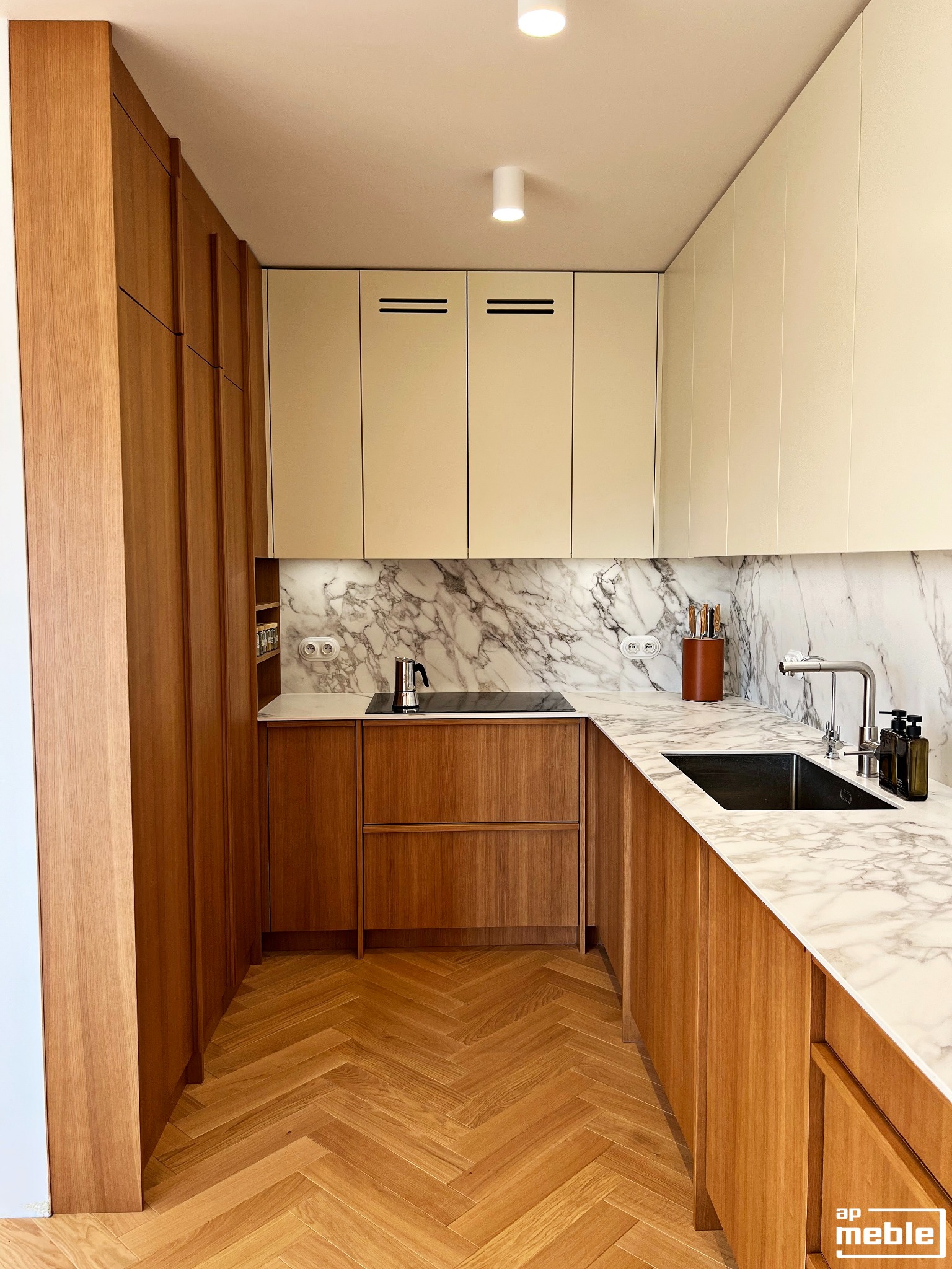 Nowoczesna kuchnia z drewnianymi frontami i blatem imitującym marmur, parkiet w jodełkę. Uwagę przykuwa minimalistyczny design i oświetlenie punktowe.