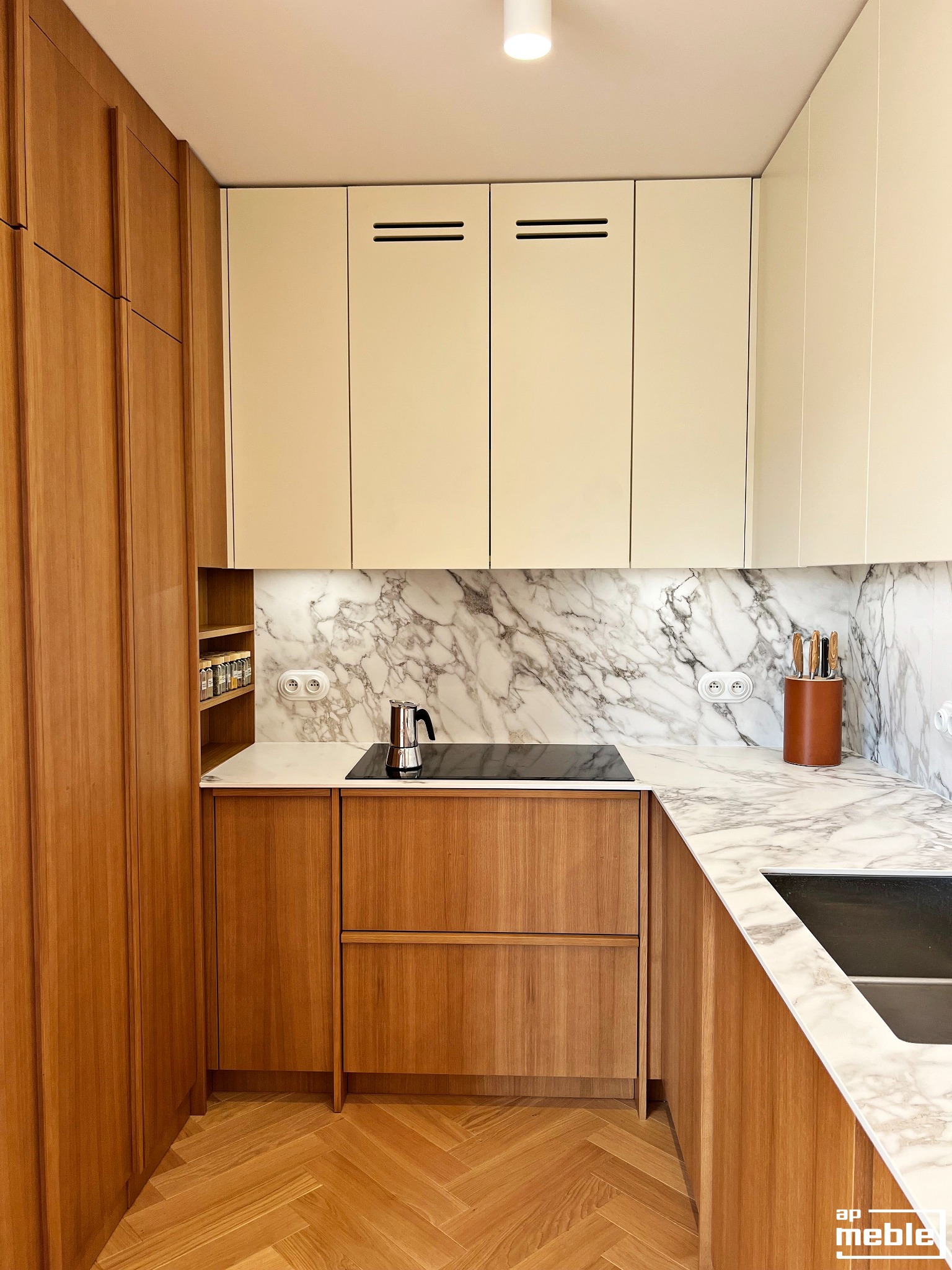 Nowoczesna kuchnia z drewnianymi frontami szafek i blatem z marmuru. Na blacie stoi srebrna kawiarka. Parkiet ułożony w jodełkę. Górne szafki w kolorze kremowym.
