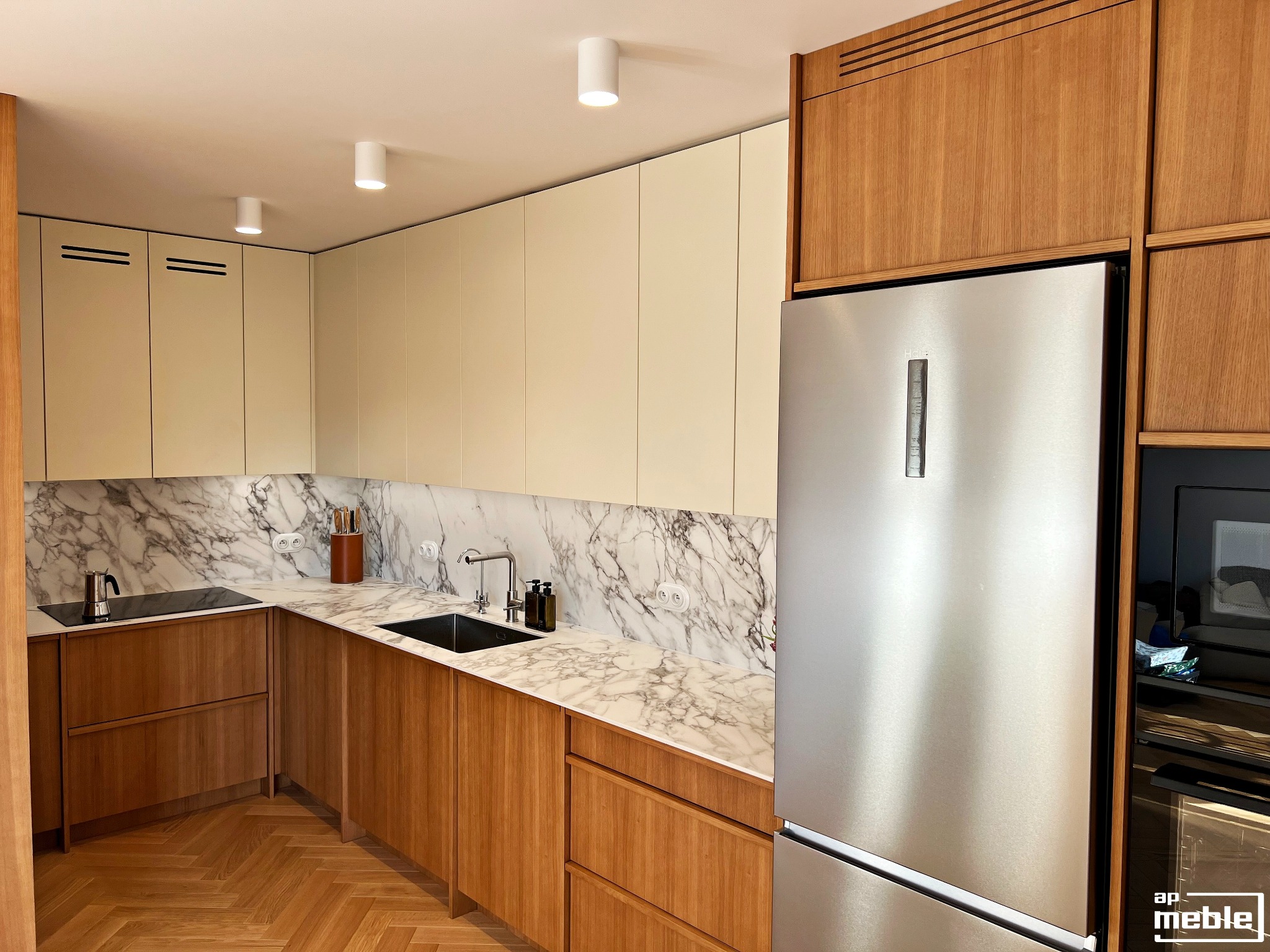 Nowoczesna kuchnia z drewnianymi frontami szafek, marmurowym blatem i srebrną lodówką. Parkiet w jodełkę na podłodze. Minimalistyczne oświetlenie sufitowe.