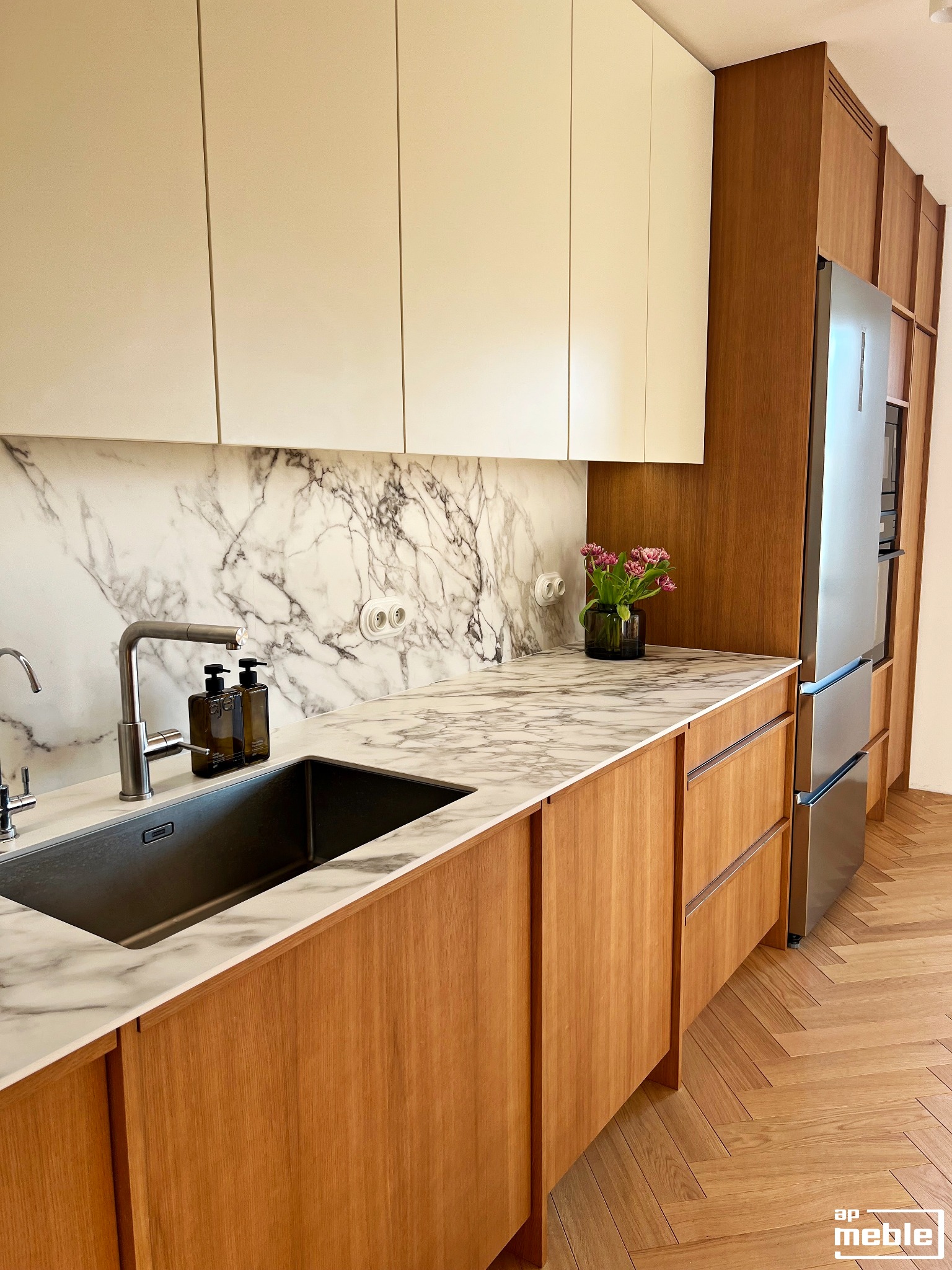 Nowoczesna kuchnia z drewnianymi frontami, marmurowym blatem i backsplashiem. Widoczny zlew, bateria i kwiaty w wazonie. Uwagę przykuwa parkiet ułożony w jodełkę.