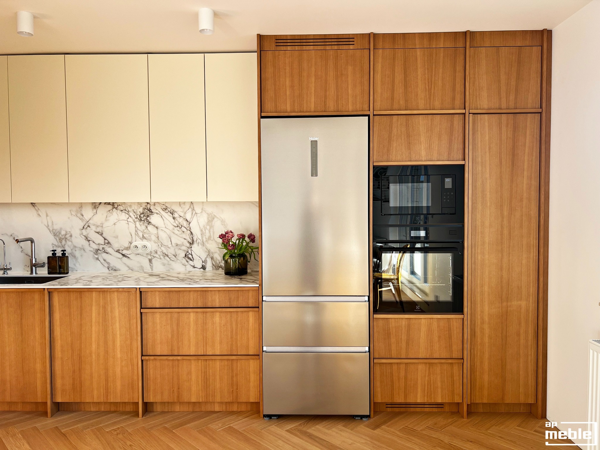 Nowoczesna kuchnia z drewnianymi frontami i marmurowym blatem. Srebrna lodówka Haier w zabudowie. Parkiet w jodełkę. Minimalistyczne oświetlenie sufitowe.