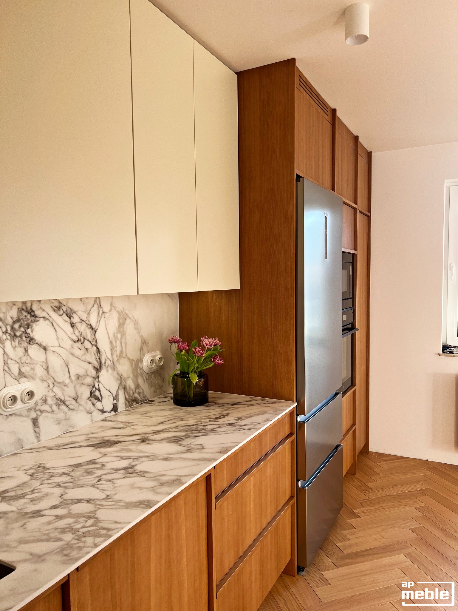 Nowoczesna kuchnia z marmurowym blatem i drewnianymi frontami, uzupełniona o kwiaty w wazonie. Widoczna zabudowa lodówki i piekarnika, parkiet w jodełkę na podłodze.