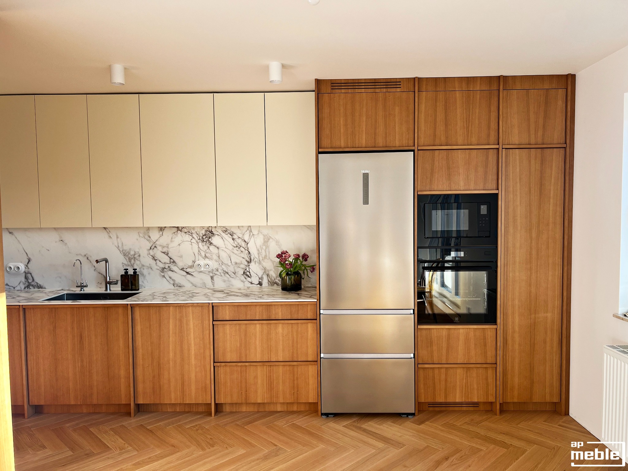 Nowoczesna kuchnia z drewnianymi frontami, marmurowym blatem i sprzętem AGD w zabudowie. Parkiet w jodełkę na podłodze. Minimalistyczne oświetlenie sufitowe.